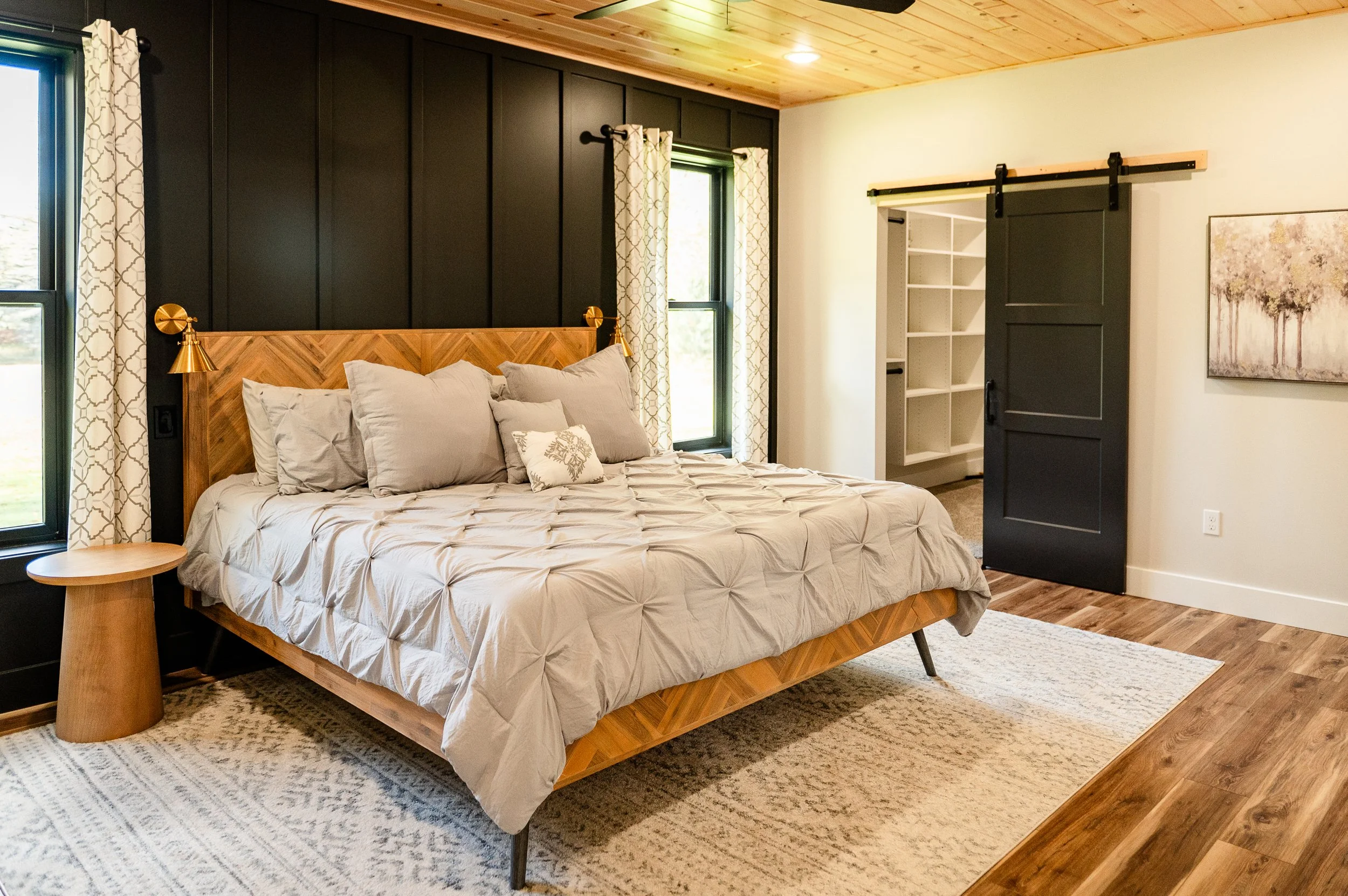 A bedroom with a wooden bed frame, beige bedding, and multiple pillows. Black paneled wall behind the bed with white patterned curtains on windows, and a sliding barn door on the right. Light wood ceiling and a floral painting on the wall.
