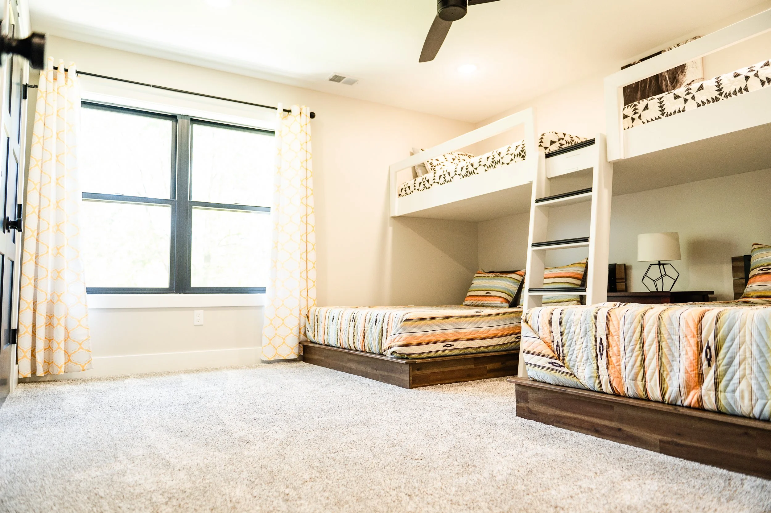 Room with two bunk beds with black and white geometric bedding, a window with white curtains, and a beige carpet.