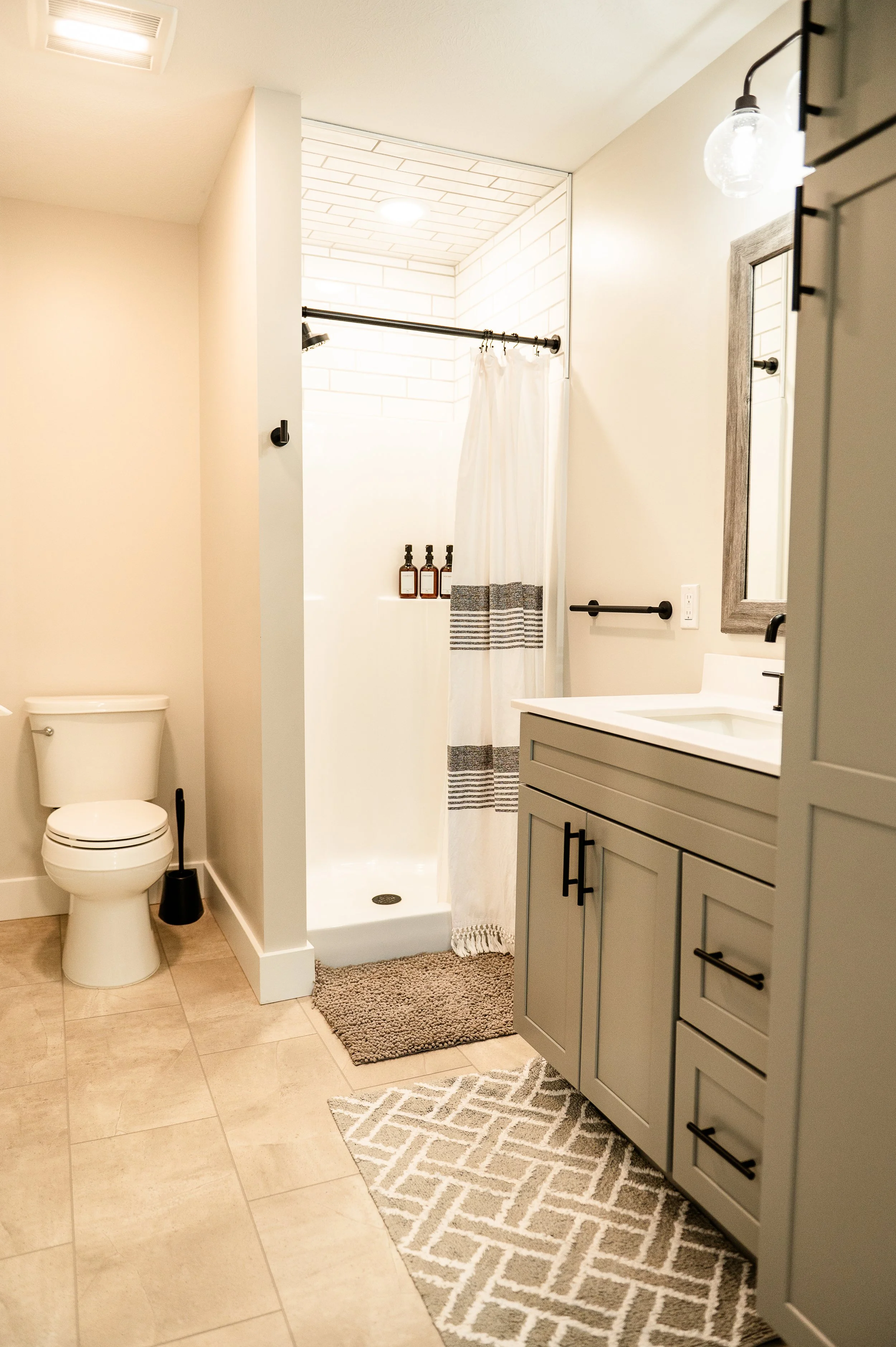 A bathroom with a toilet, a shower with white subway tile, bottles on the shower shelf, a beige and white striped shower curtain, a bathroom vanity with a mirror and a light fixture above, a beige rug and a patterned gray and white rug on the tiled f