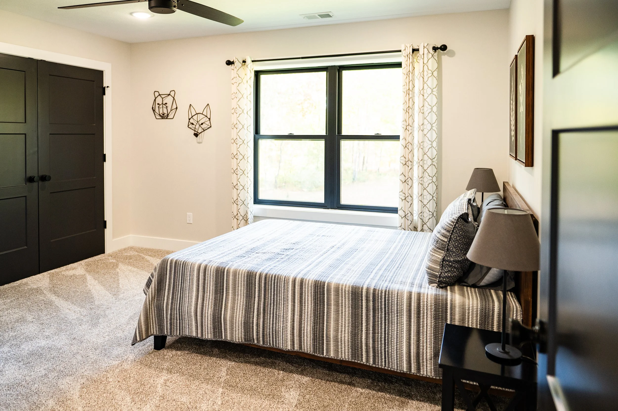 A bedroom with a bed covered in a striped bedspread, a nightstand with lamps, a window with patterned curtains, and decorative wall art of a bear and a fox.