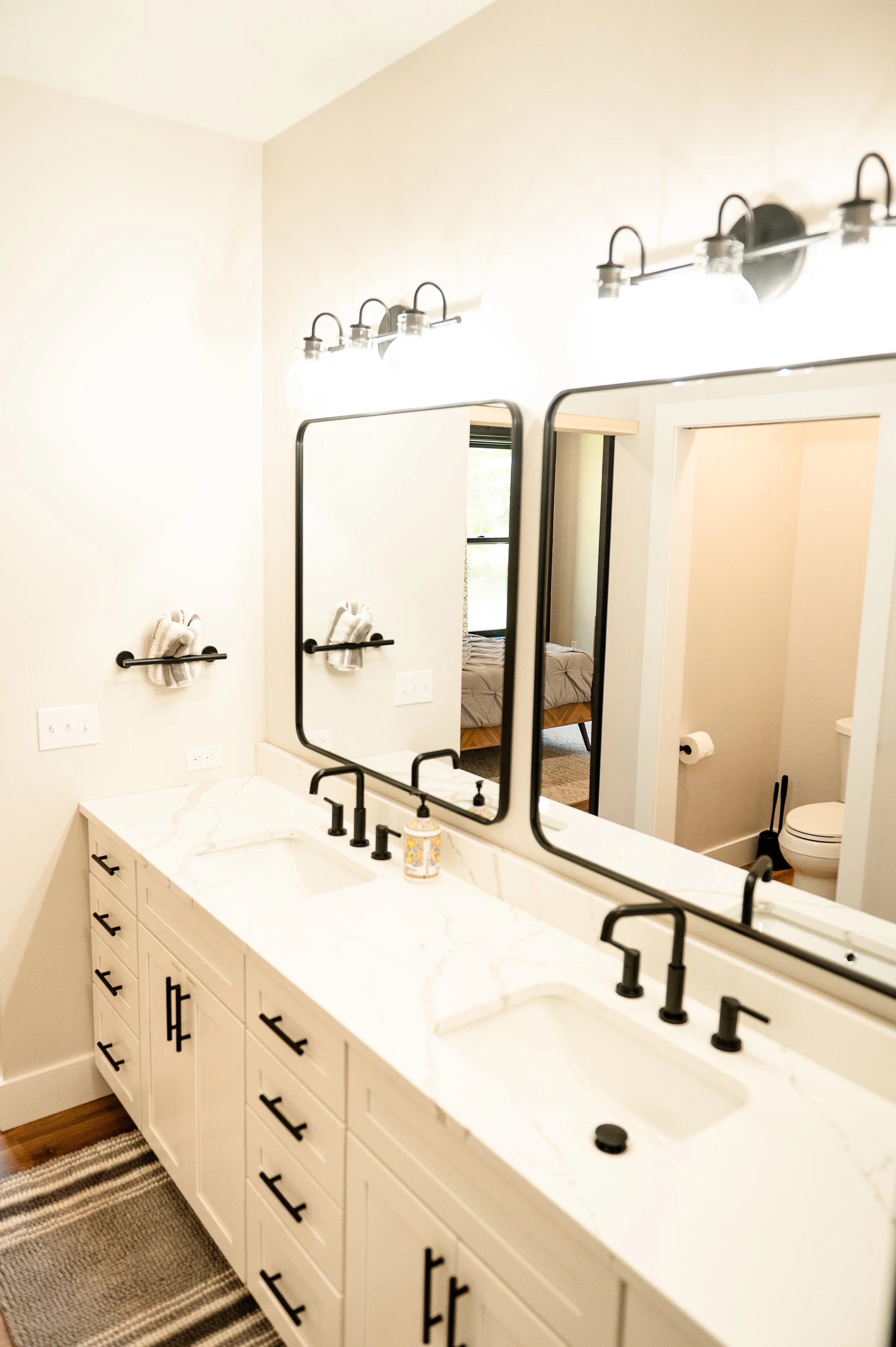 A bathroom vanity with a white marble countertop, black hardware, and two black-framed mirrors above the sinks. Light fixtures with Edison bulbs are mounted above the mirrors. The bathroom has white walls, a towel with hooks on the wall, and a view o