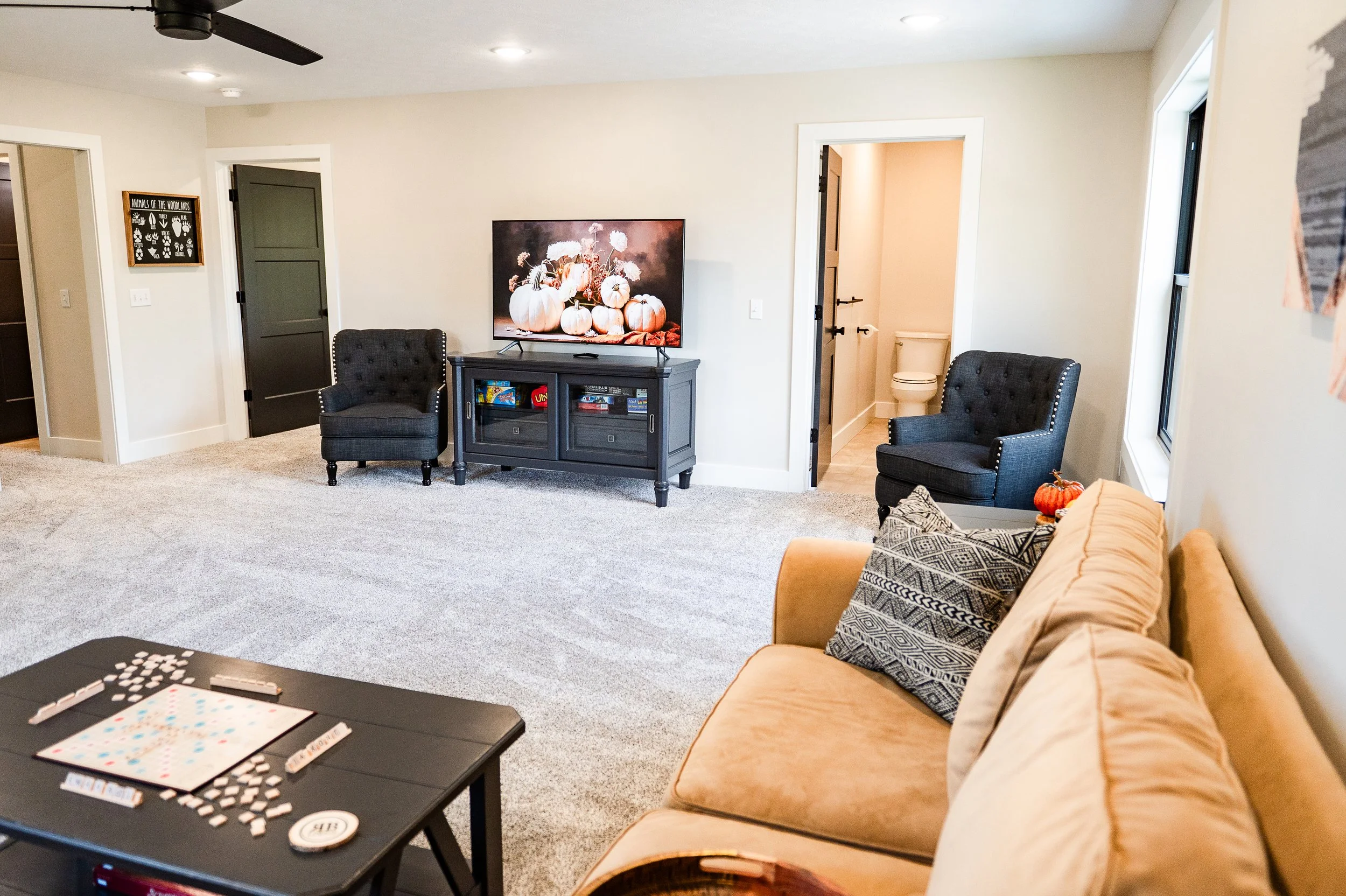 Living room with a beige sofa, black coffee table playing board games, two black armchairs, a TV with pumpkins and flowers on the screen, a dark gray door, a wall with a decorative frame, and a small bathroom visible through an open door.