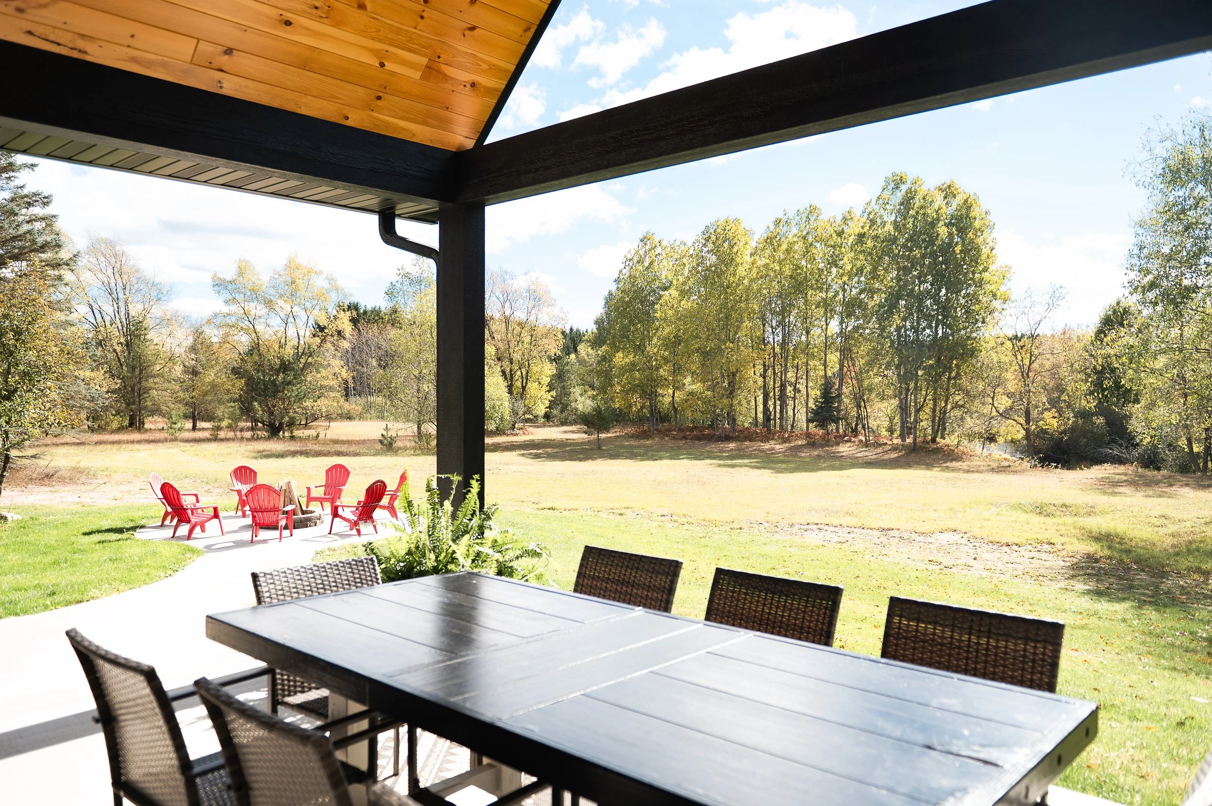 View from a covered outdoor patio with black tables and chairs, overlooking a grassy yard with red Adirondack chairs and a background of trees with green and yellow leaves under a partly cloudy sky.