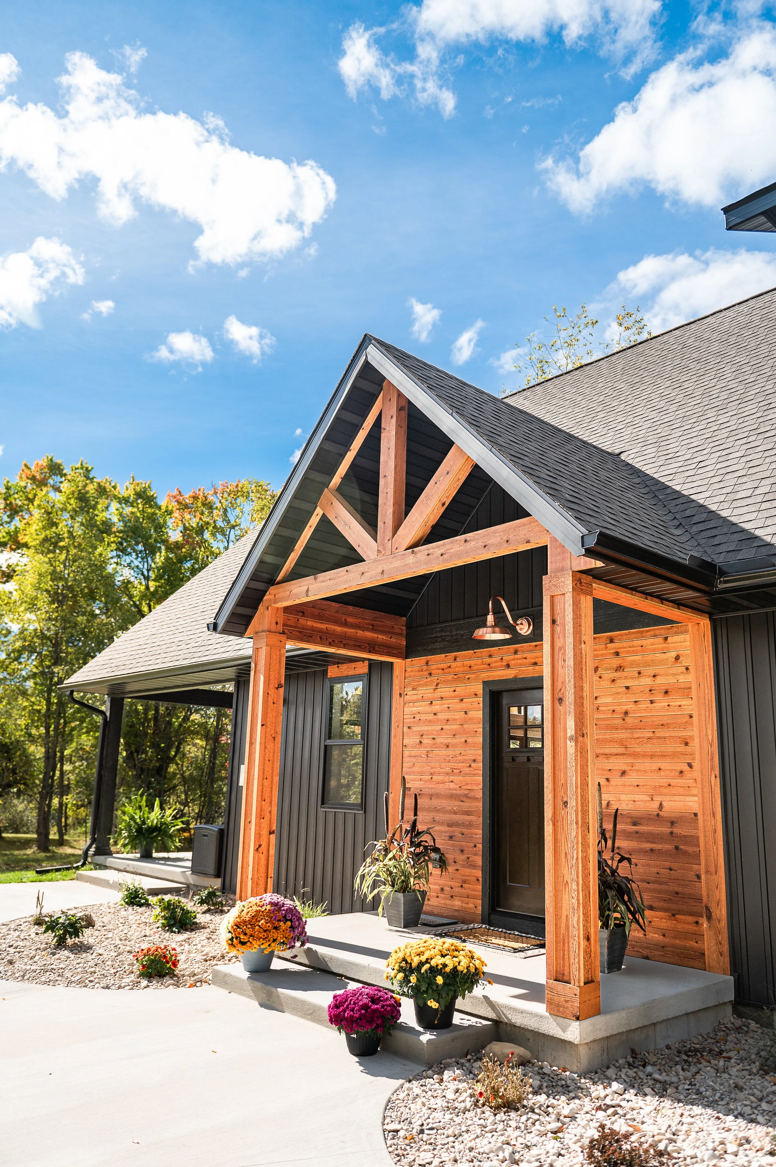 A house under construction with a black exterior and wooden accents, featuring a gabled roof and outdoor lighting, surrounded by potted flowers and landscaped yard, under a partly cloudy sky.