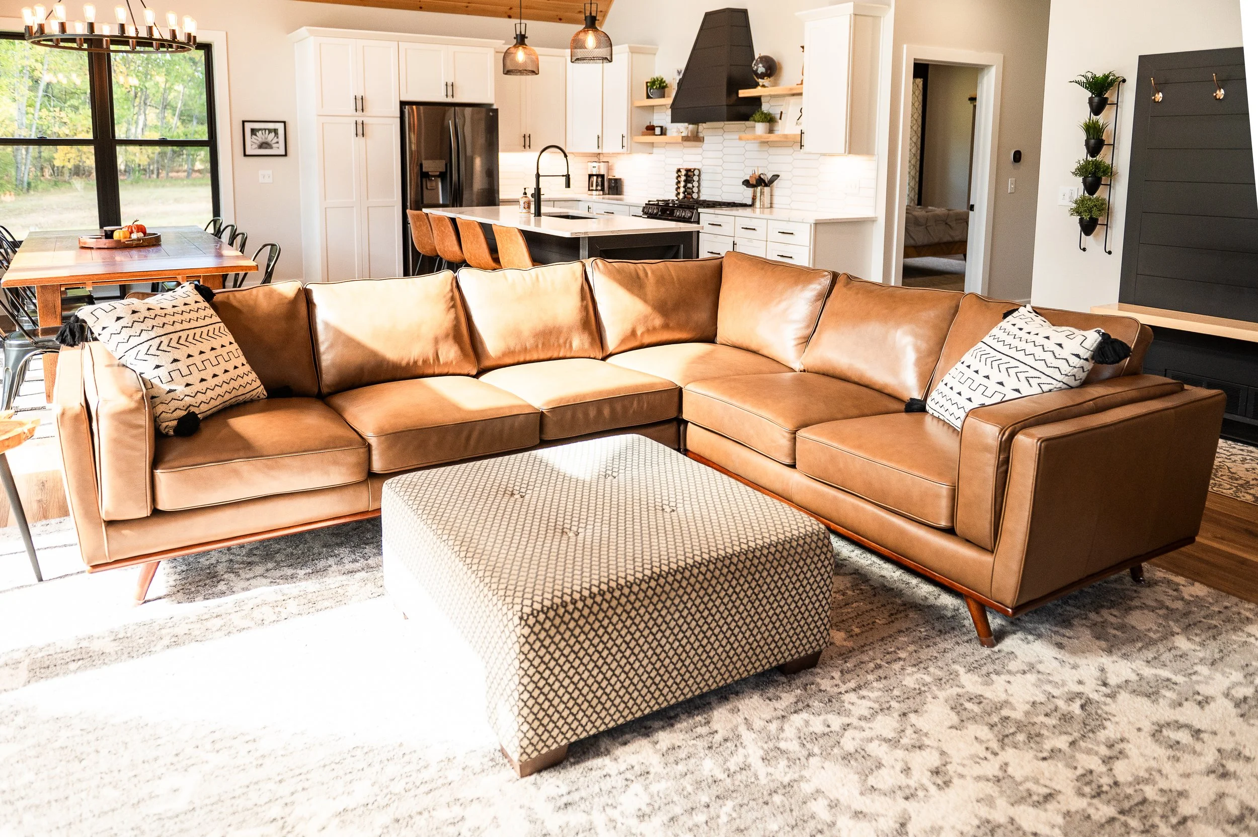 Living room and kitchen with a large leather sectional sofa, patterned ottoman, dining table with black chairs, and modern kitchen with white cabinets, black accents, and a view of trees outside.