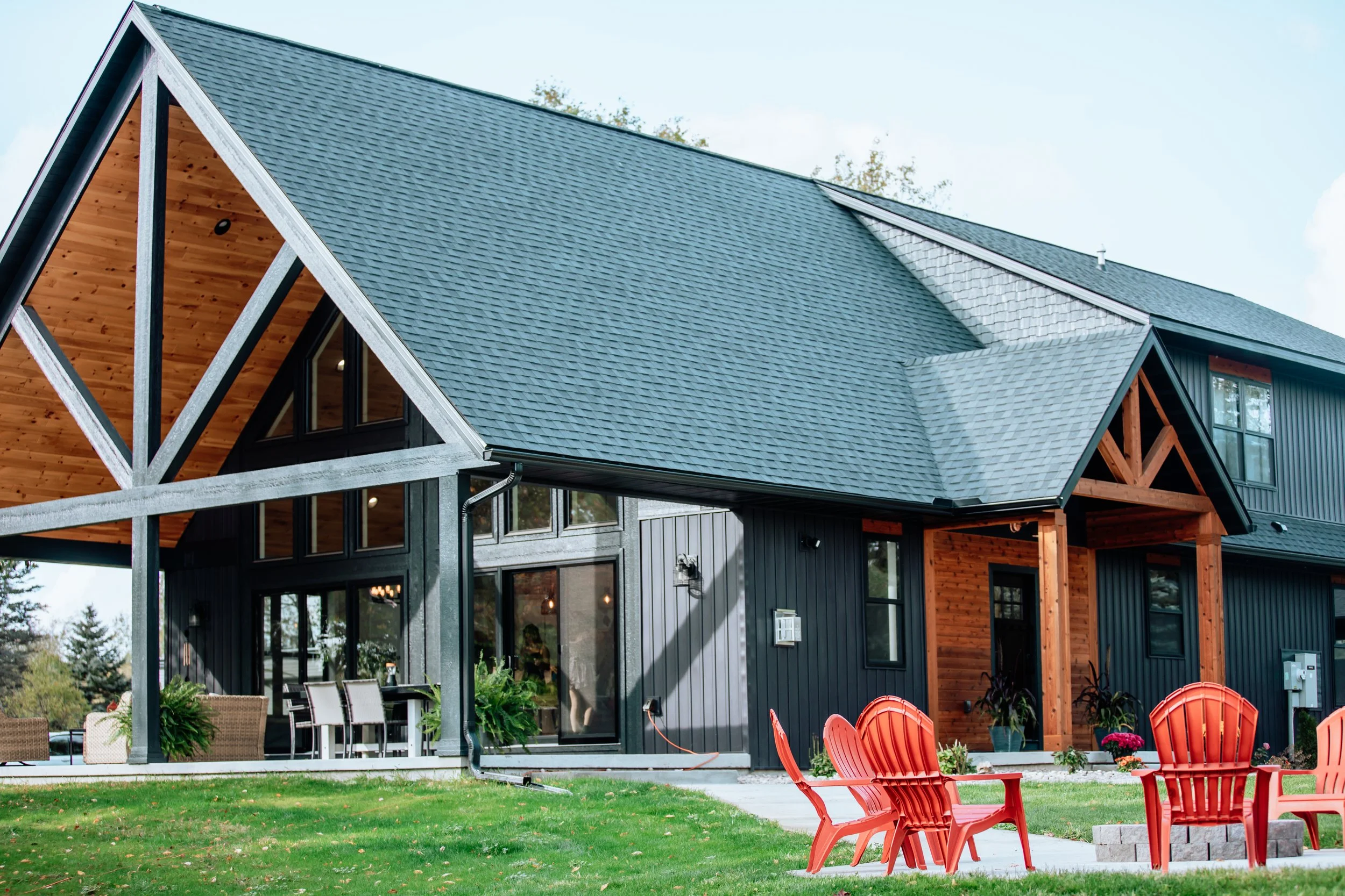 A modern house with a black exterior and large windows, featuring a steeply sloped roof with shingles. There is a wooden accent section with a triangular shape and black framing, and a spacious outdoor area with red Adirondack chairs, dining table, a