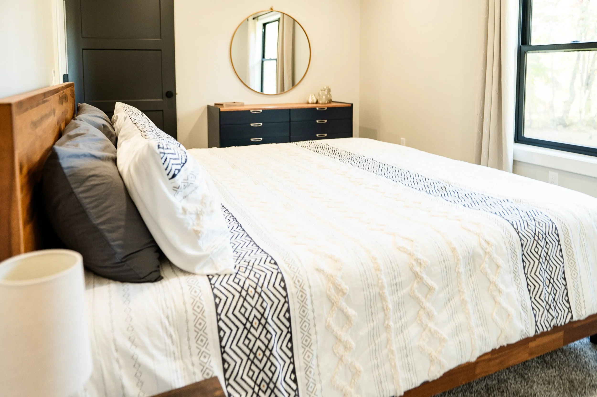 A neatly made bed with a wooden headboard, with decorative pillows and a patterned white bedspread, in front of a black dresser with gold handles and a round mirror above it, in a bedroom with large windows and white walls.