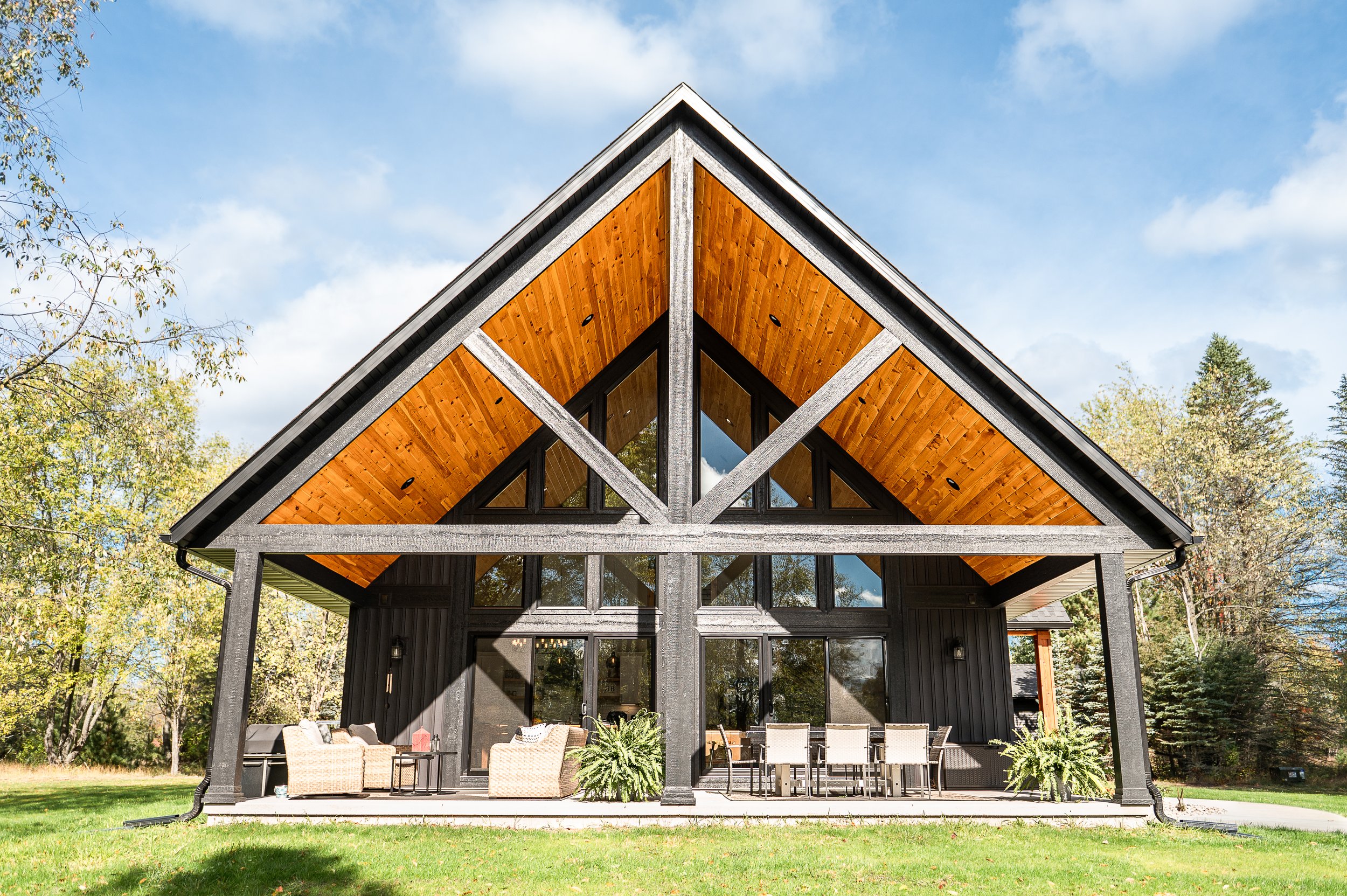 Modern house with black exterior, large glass windows, and a prominent overhanging roof with wooden underside, situated in a wooded area on a sunny day.