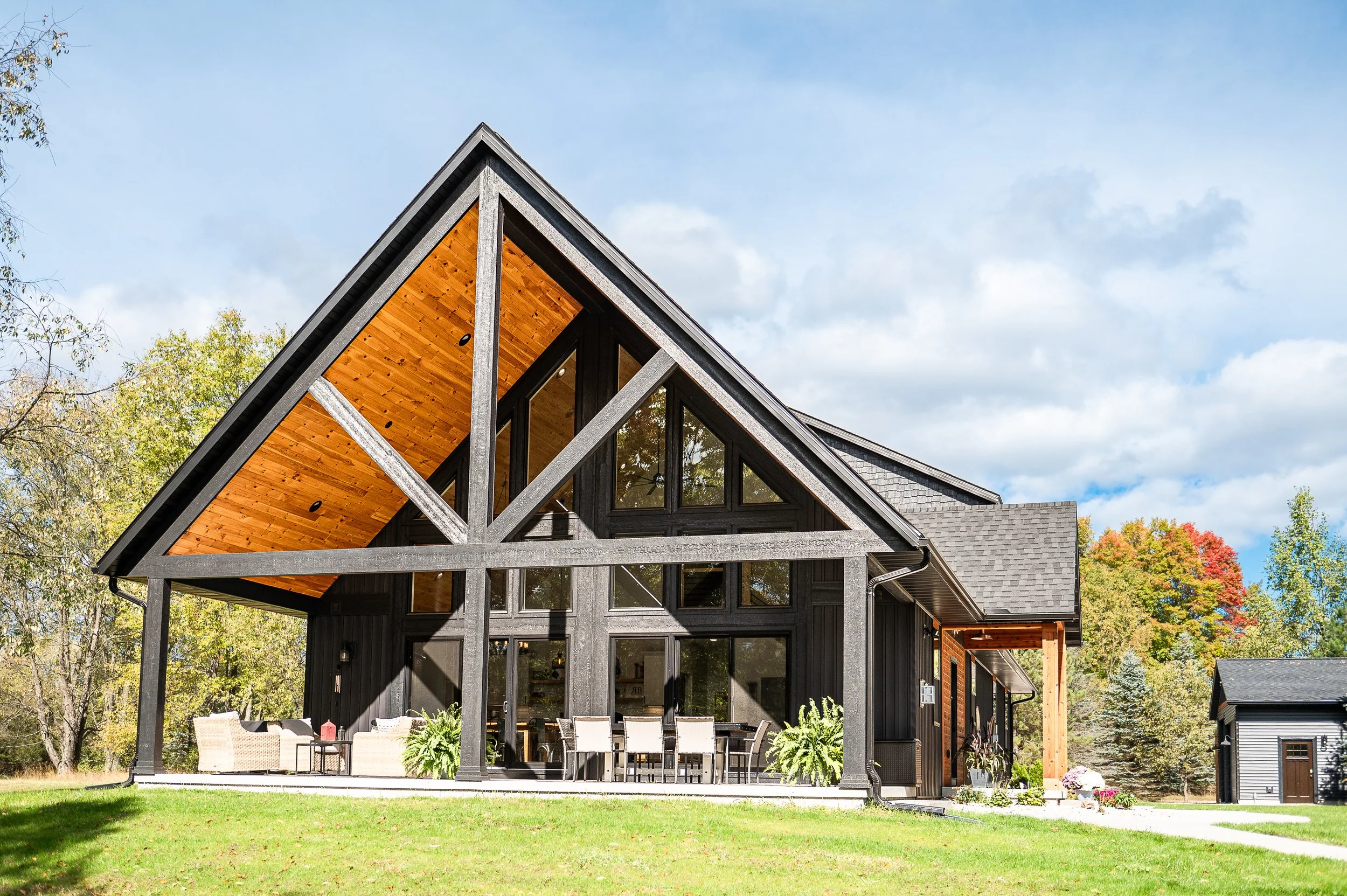 Modern black house with large windows, exposed wooden roof, and outdoor patio with dining furniture, set on a grassy lawn with trees and a blue sky in the background.
