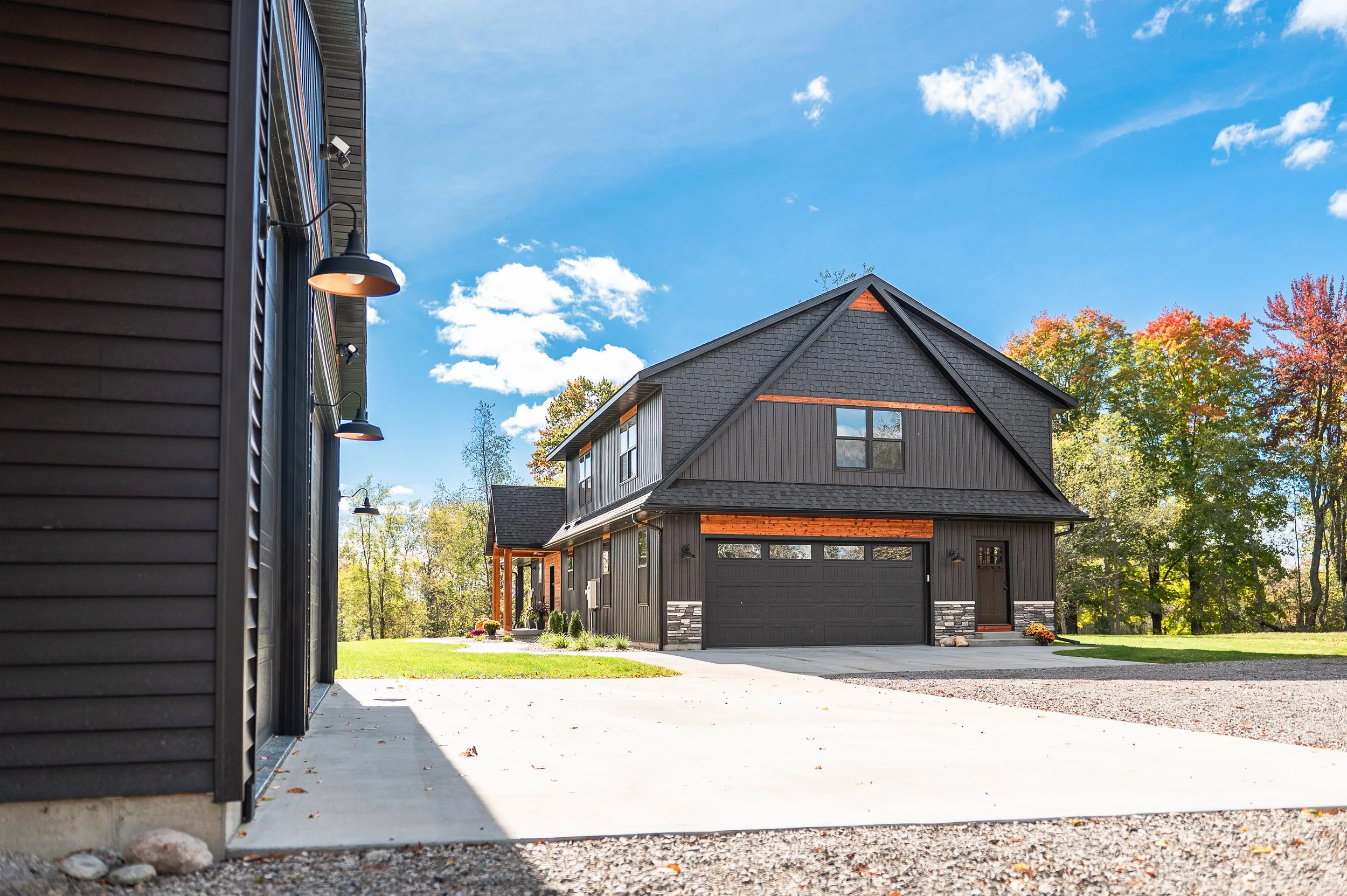 View of a modern house with a dark exterior, black garage door, and stone accents, surrounded by trees with fall foliage under a bright blue sky with some clouds.