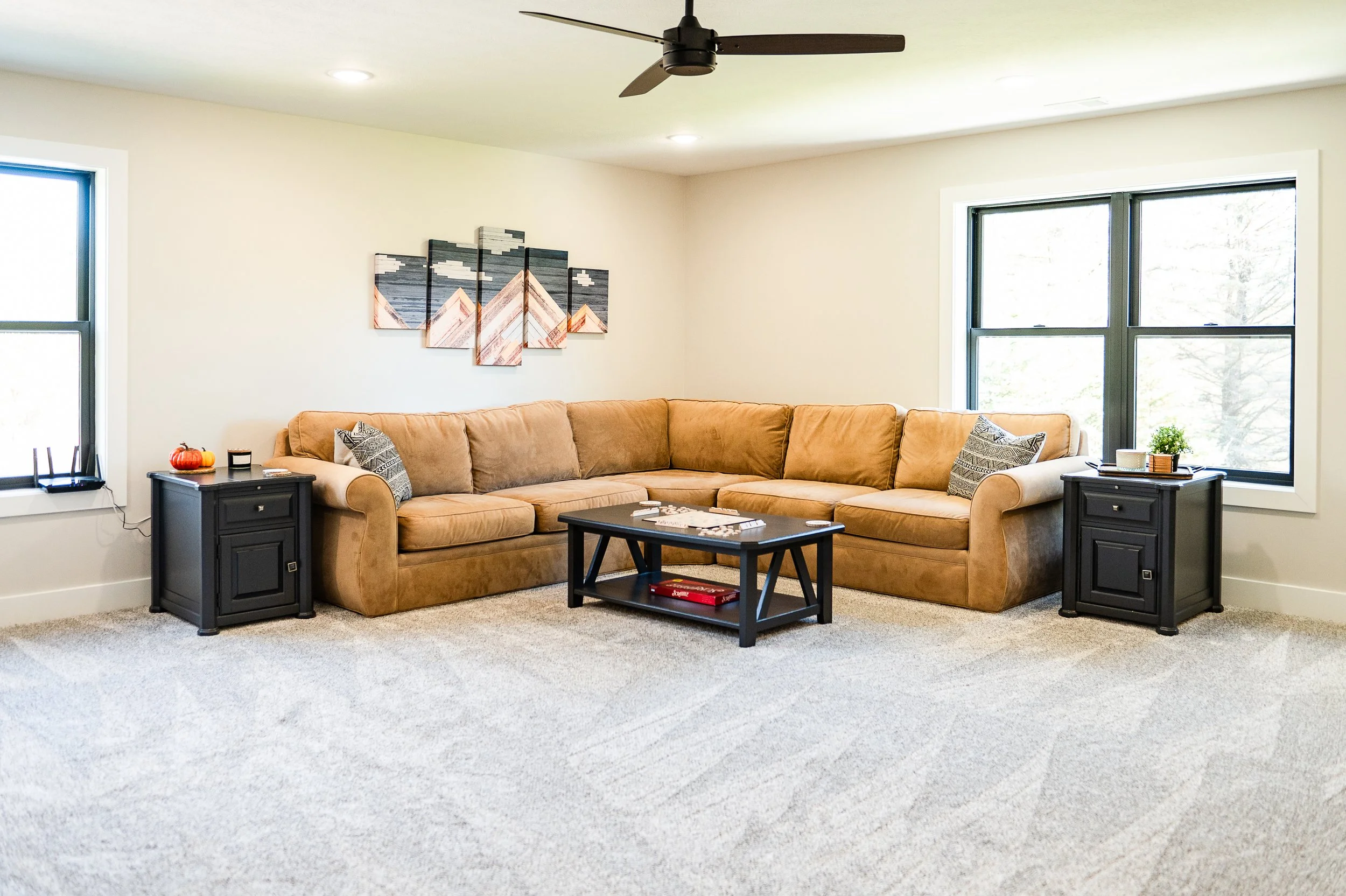 A living room with a tan sectional sofa, black side tables, and large windows with forest view. A wall art piece with geometric mountain design and a small pumpkin decoration are on one side table. There is a ceiling fan and recessed lighting.