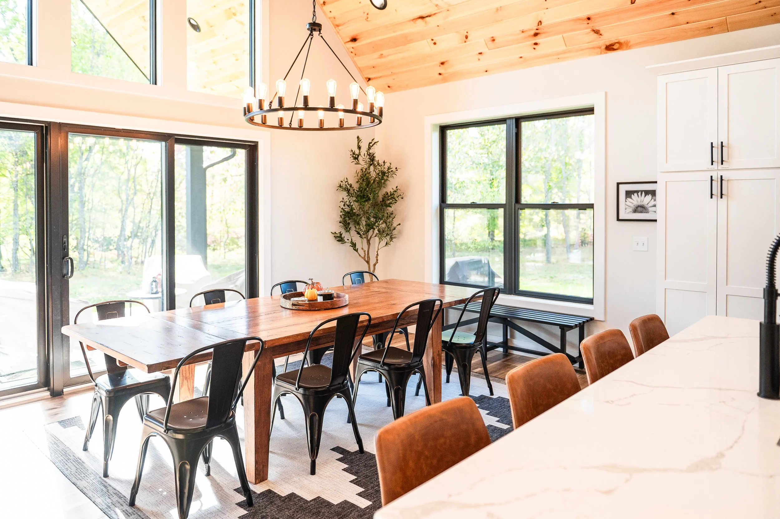 The image shows a bright, modern dining room with a large wooden dining table surrounded by black metal chairs. There are large windows letting in natural light, a chandelier hanging from the wooden ceiling, and a decorative tray with pumpkins on the