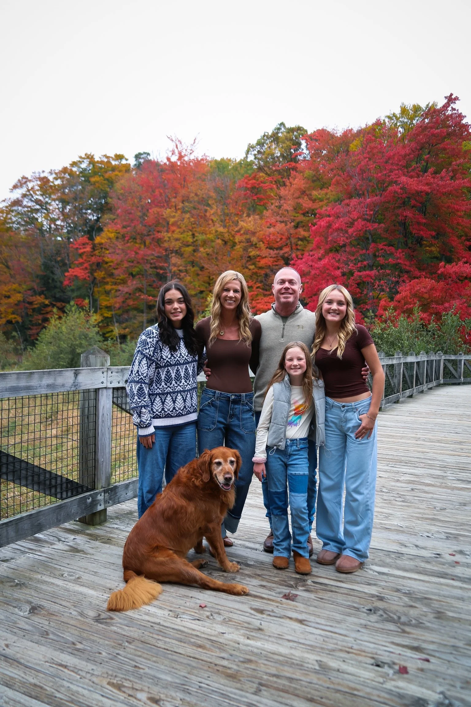 A family of six, including an adult man, adult women, three young girls, and a dog, standing on a wooden bridge with fall foliage in the background.