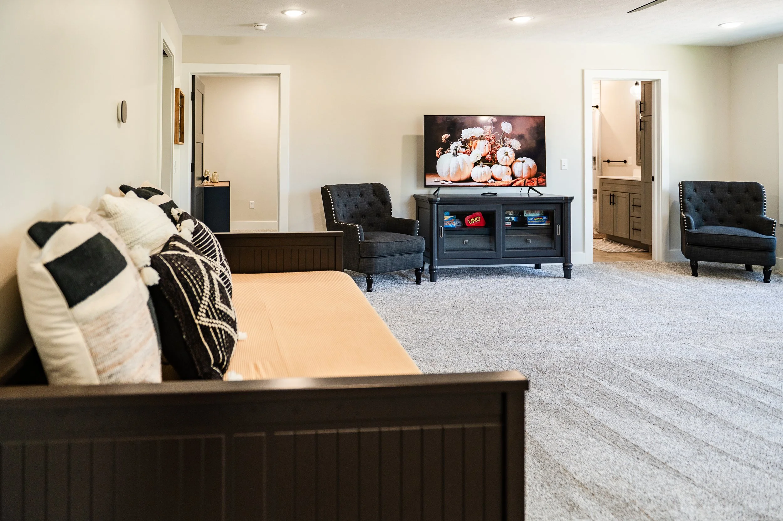 Living room with a beige sofa with black and white pillows, a black armchair, TV on a black stand featuring pumpkins and flowers on the screen, and an open doorway to a bathroom.