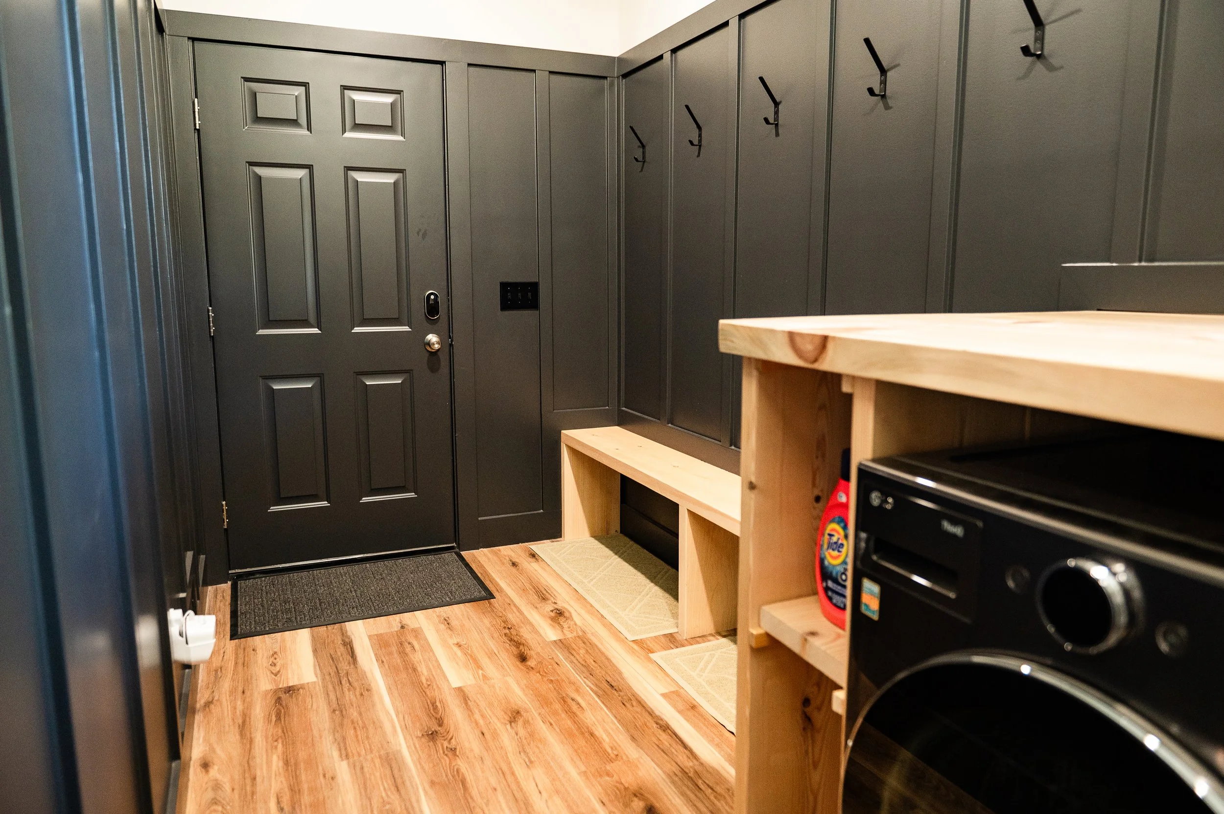 A small room with a black front door and black hooks on the wall. There are two small beige rugs on the wooden floor, and a partially visible wooden countertop with a box of Tide laundry detergent and a black appliance.