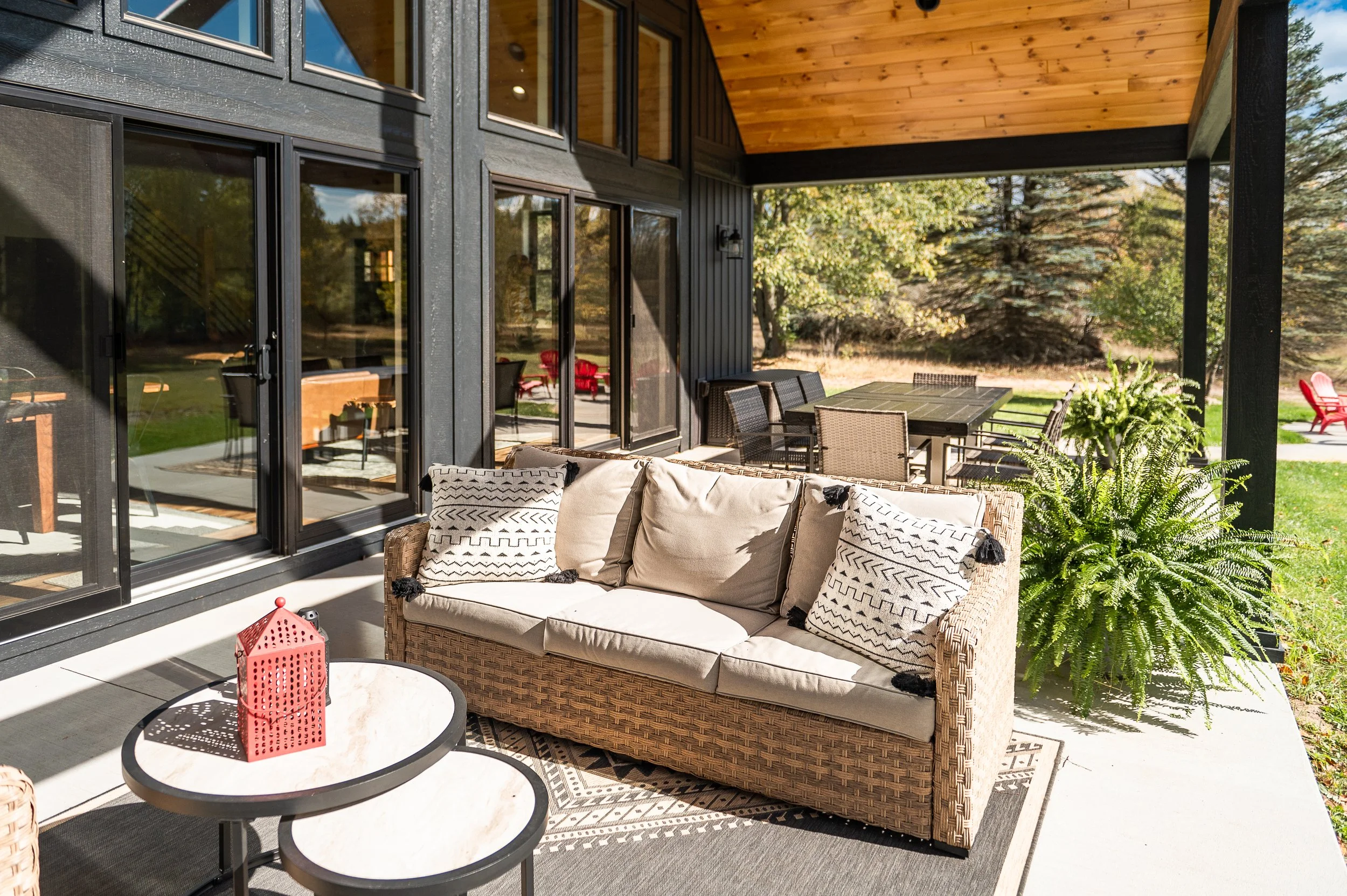 Outdoor patio with wicker sofa with beige cushions, black and white pillows, a round marble-topped coffee table with red lantern, and a black dining table with chairs underneath a wooden ceiling, surrounded by trees.