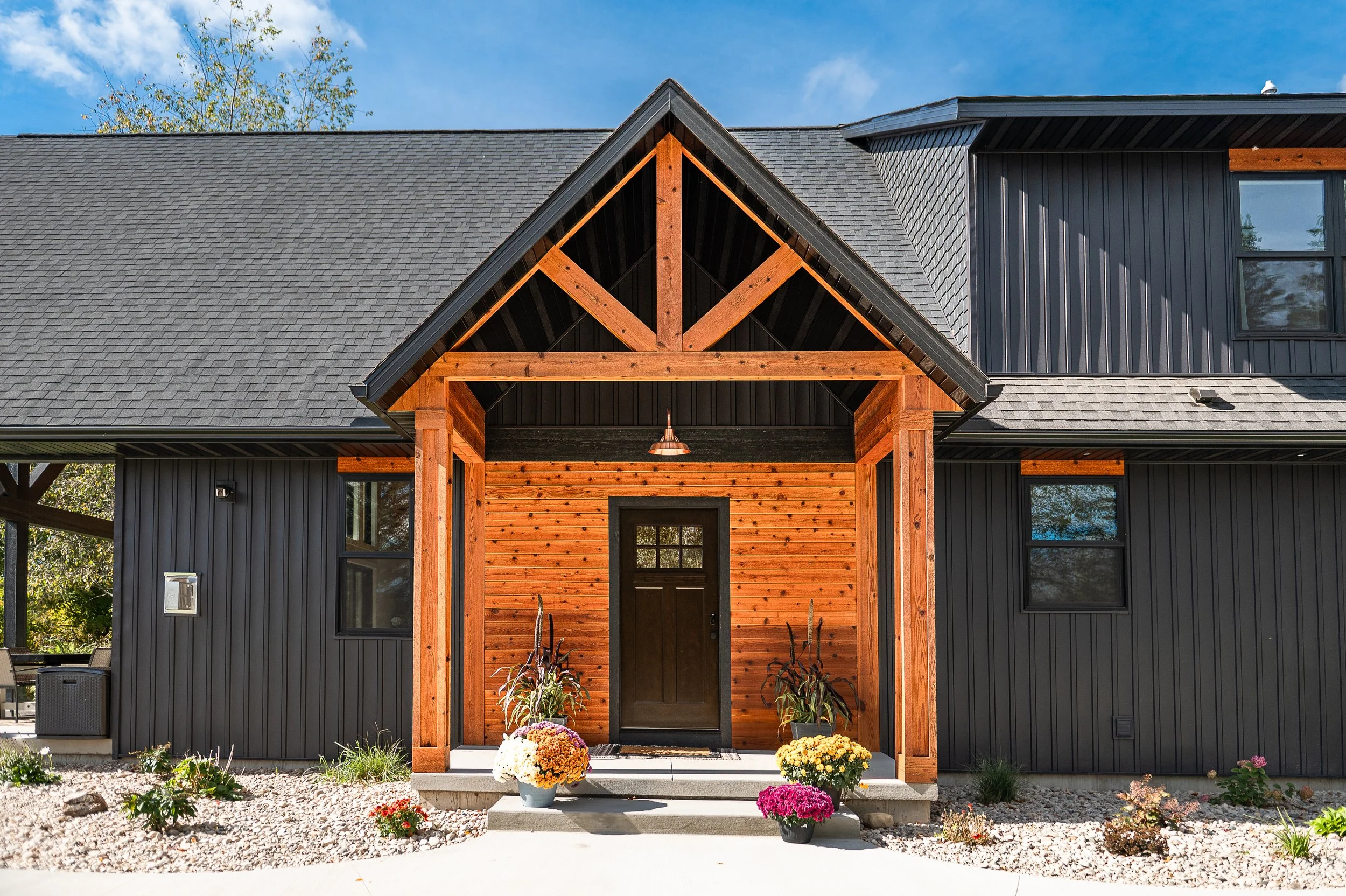 Front view of a modern house with a gable roof, black siding, and a covered porch with wooden beams, decorated with potted flowers on the steps.