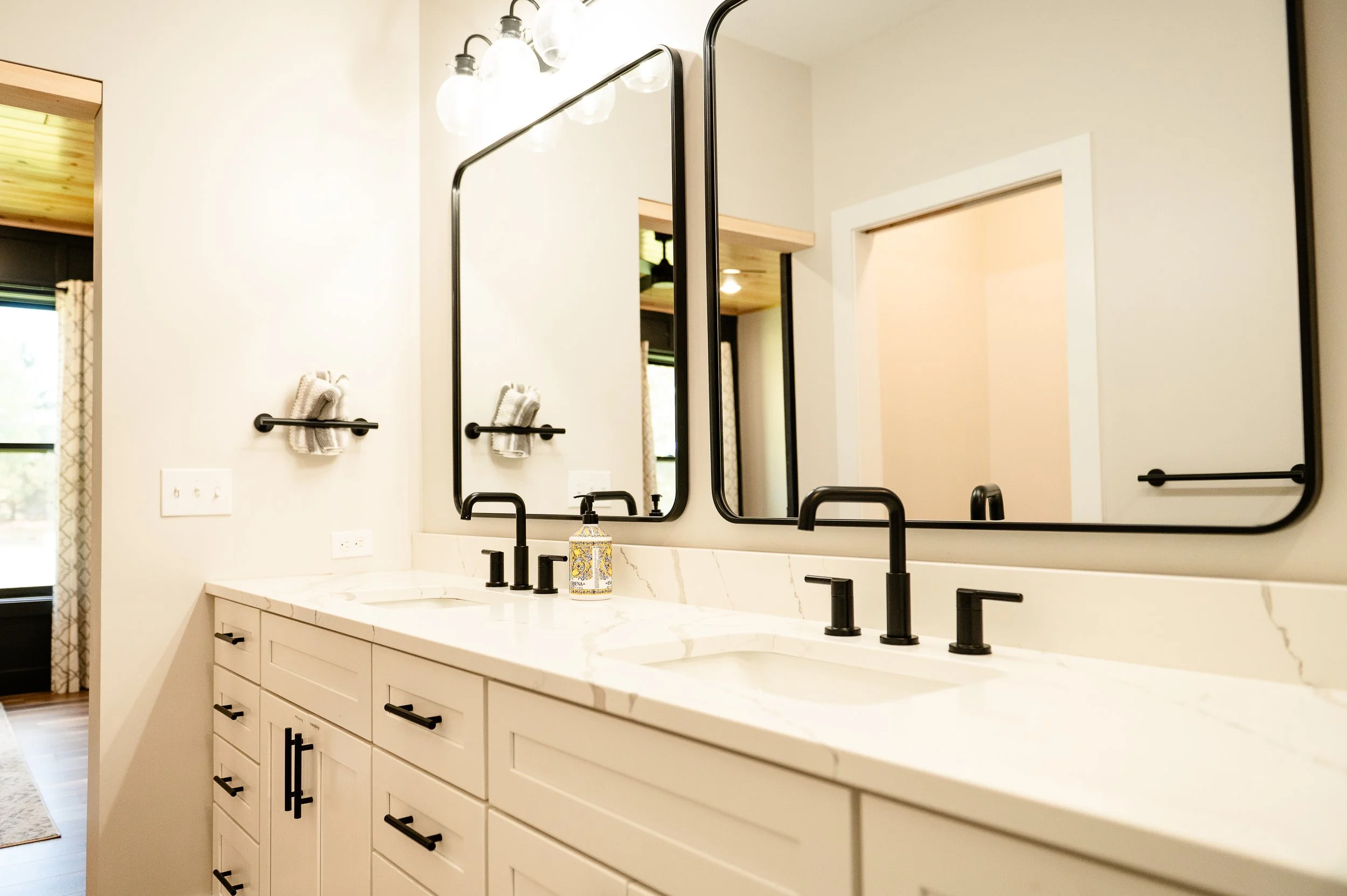 Modern bathroom vanity with white cabinetry, black handles, black faucets, a marble countertop, two large mirrors, and a wall-mounted light fixture.
