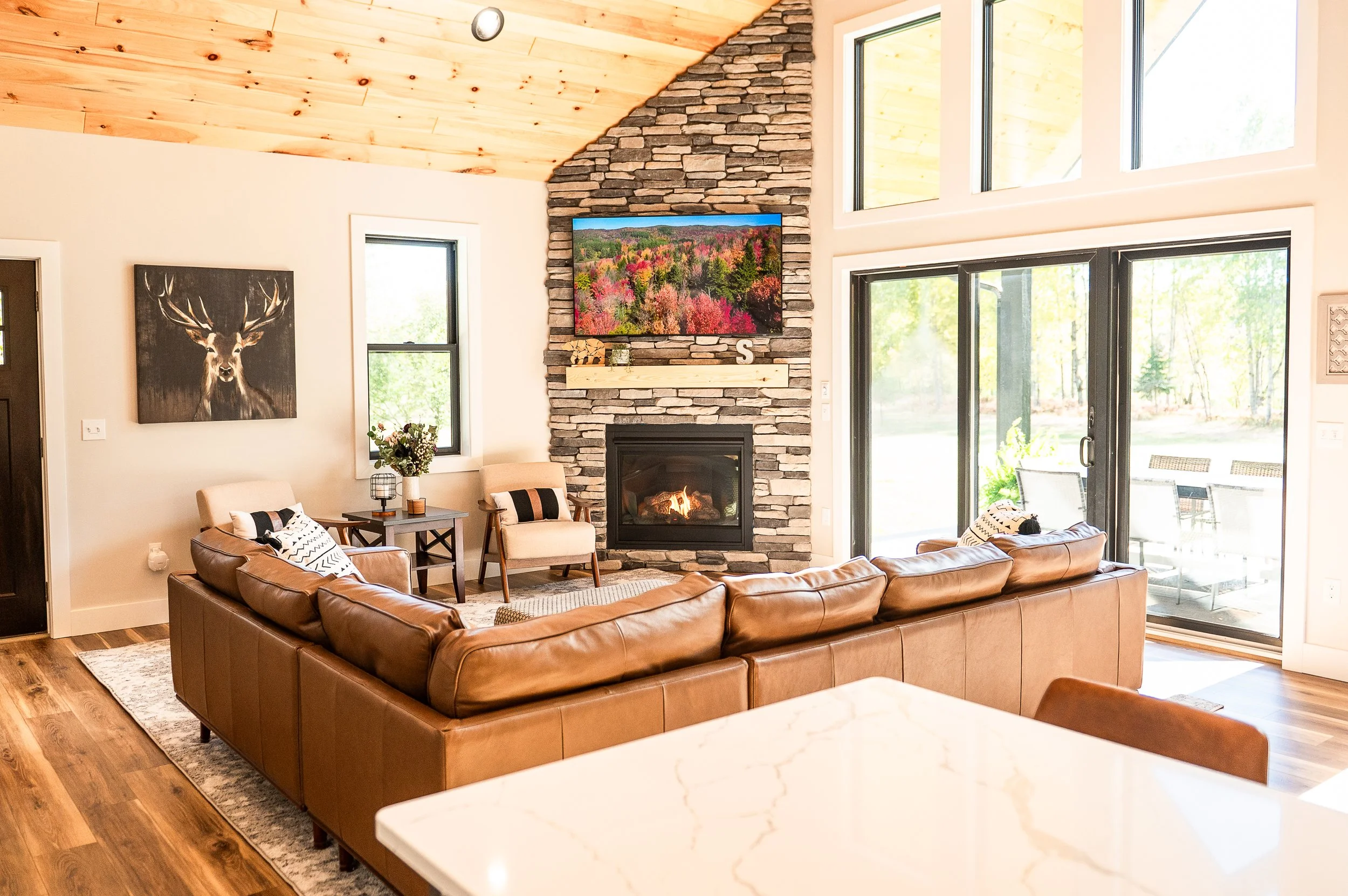 Living room with a brown leather sofa, two beige chairs, a stone fireplace with a flat-screen TV mounted above, a picture of a deer on the wall, and large windows showing outdoor greenery.