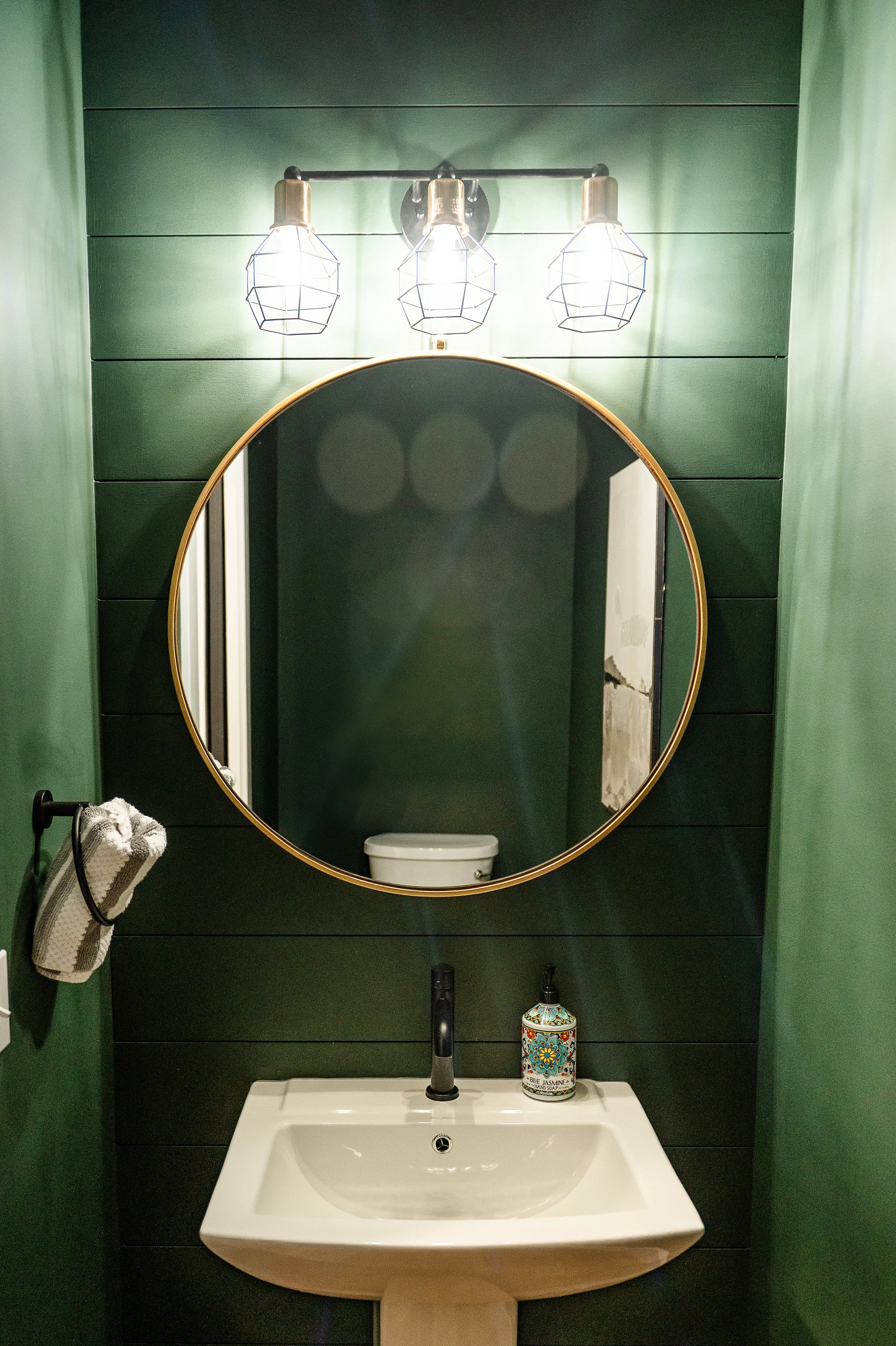 A bathroom vanity area with a wall-mounted oval mirror with a gold frame. Above the mirror are three exposed bulb light fixtures with geometric wire shades. The wall behind the mirror is covered in dark green horizontal panels. To the left of the mir