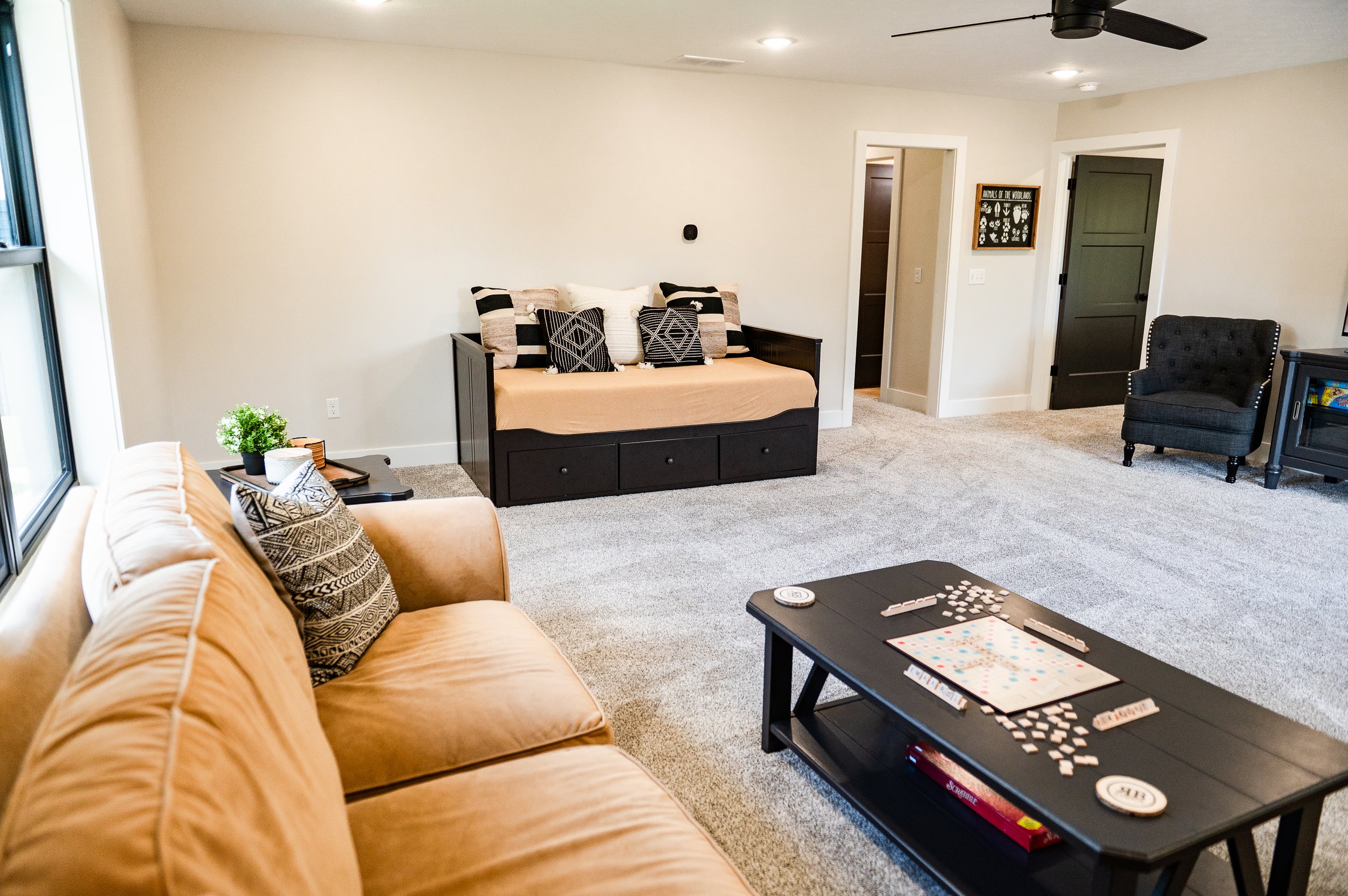 Living room with beige couch, black table with board game, and additional seating, beige walls, carpeted floor, large window.