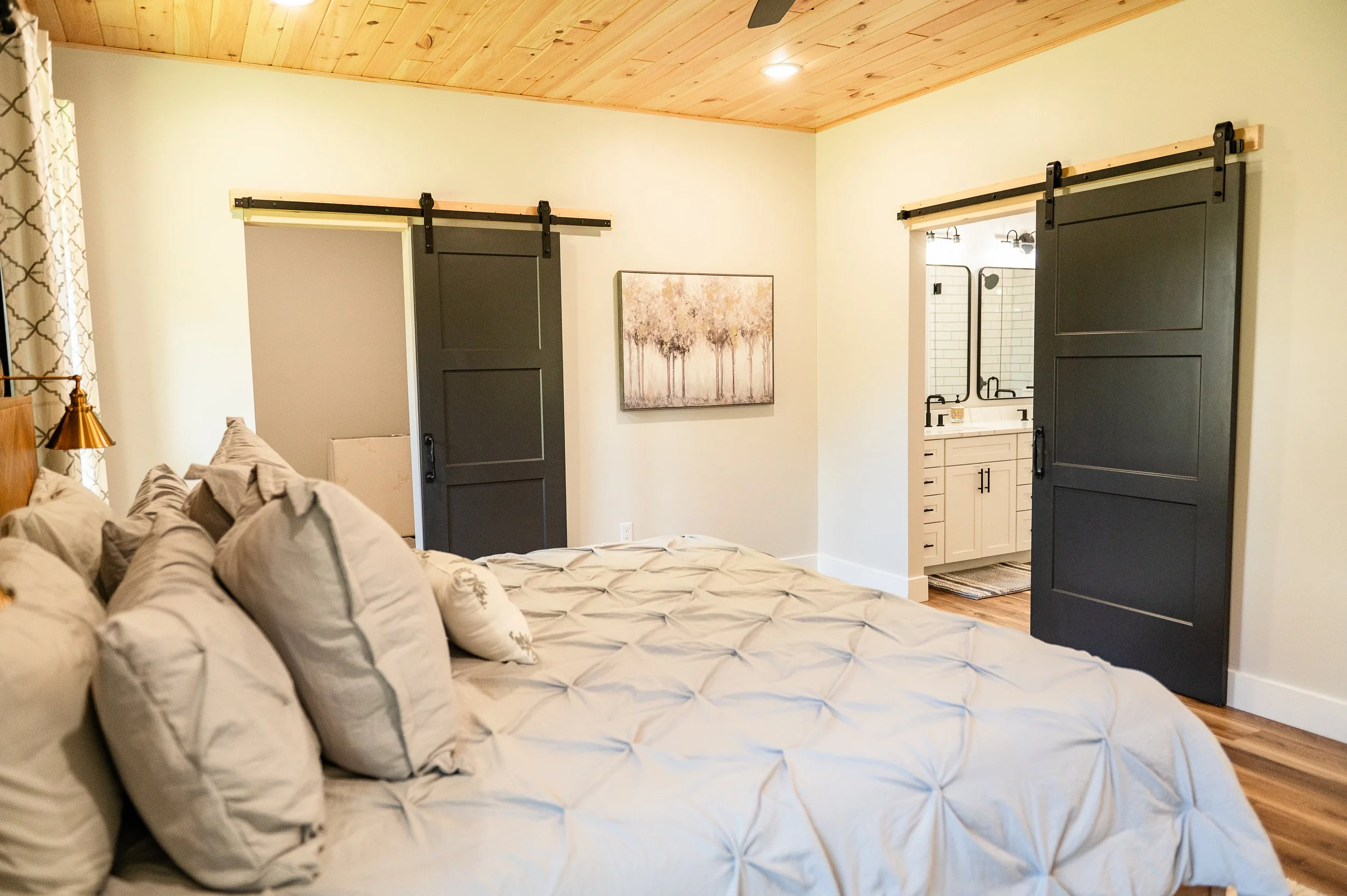 Bedroom with a bed featuring beige and white patterned bedding and pillows, yellow walls, wooden ceiling, and a sliding barn door leading to a bathroom with white tile walls and a white vanity.