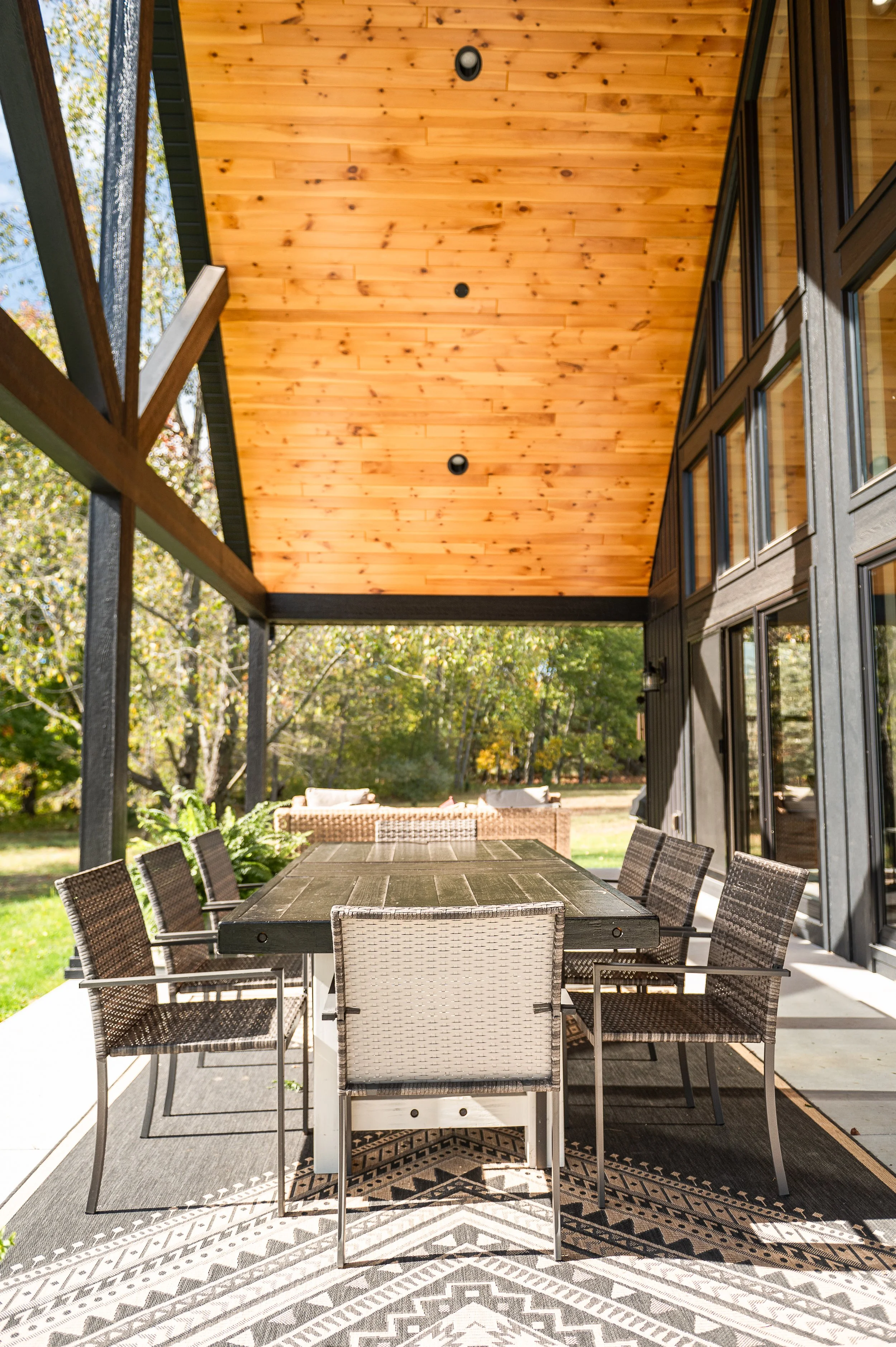 Outdoor patio with a large rectangular table surrounded by chairs, covered by a wooden ceiling, with trees in the background and a patterned outdoor rug underneath the table.