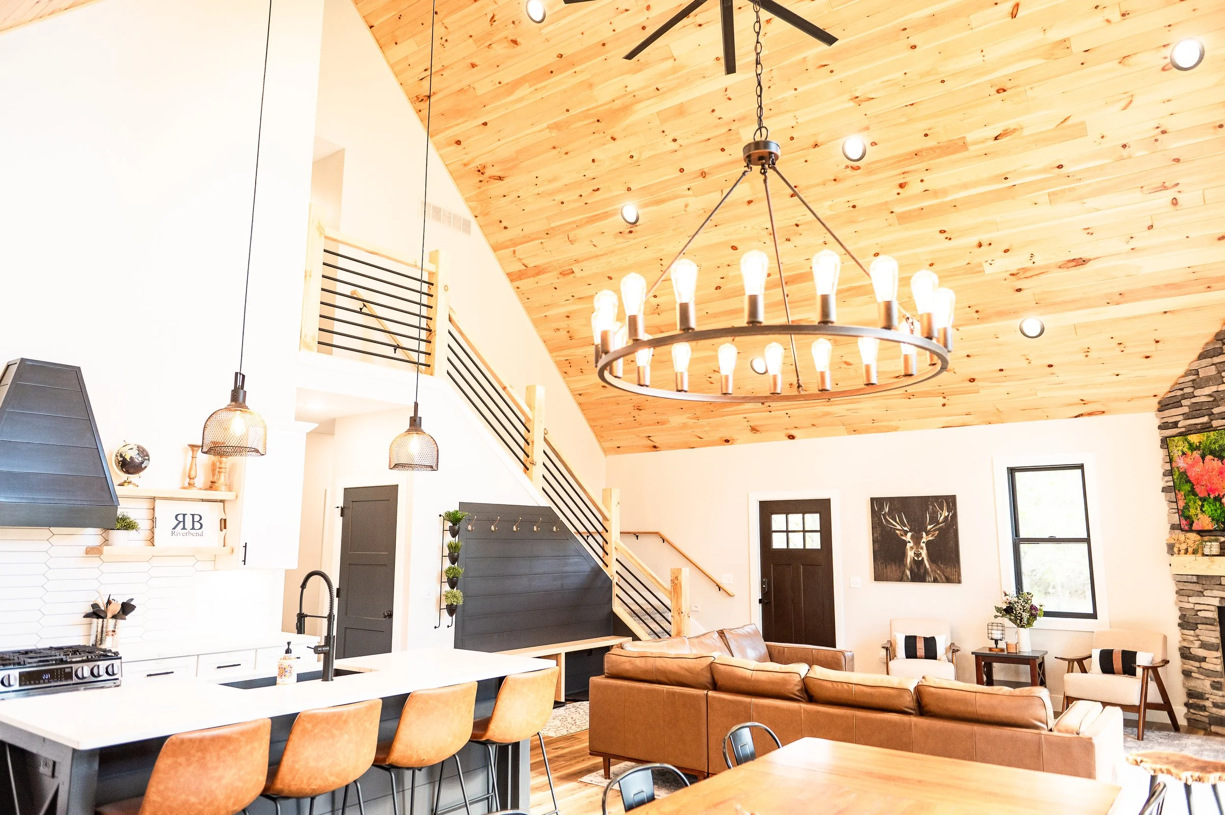 Open-concept living space with a vaulted wood ceiling, large chandelier, and modern decor. Kitchen island with beige chairs, and a cozy seating area with brown leather couches. Decor includes potted plants, wall art, and warm lighting.