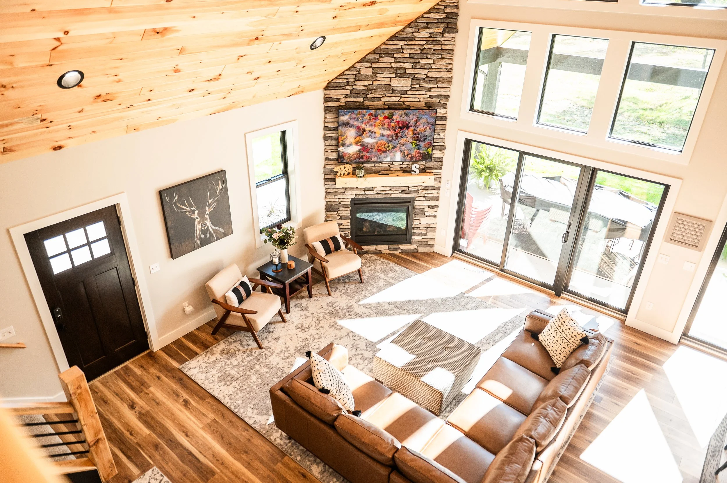 Living room with high sloped wooden ceiling, large windows, a stone fireplace with a colorful artwork above, a leather sofa, armchairs, a rug, and outdoor view.