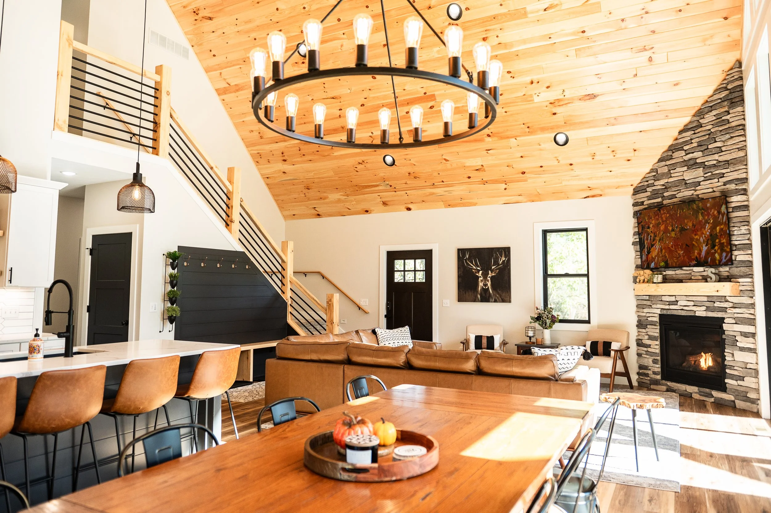 Open-concept living room with a wooden vaulted ceiling, a large chandelier, stone fireplace, and modern decor including a painting of a deer and autumn leaves.