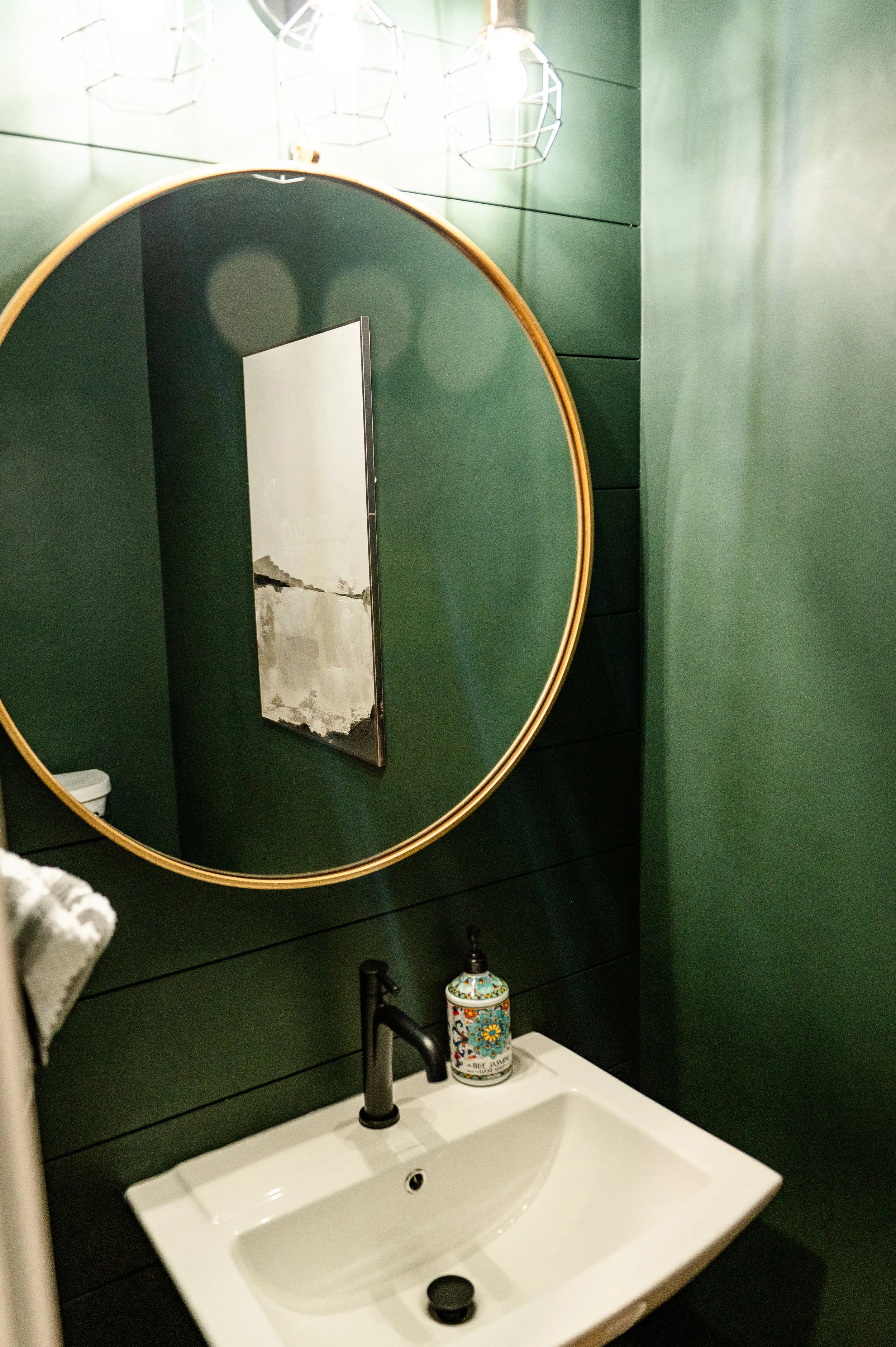 Bathroom mirror reflecting a wall with three modern geometric pendant lights, green wall paneling, a white sink with a black faucet, a decorative soap dispenser, and a gray towel hanging on a hook.