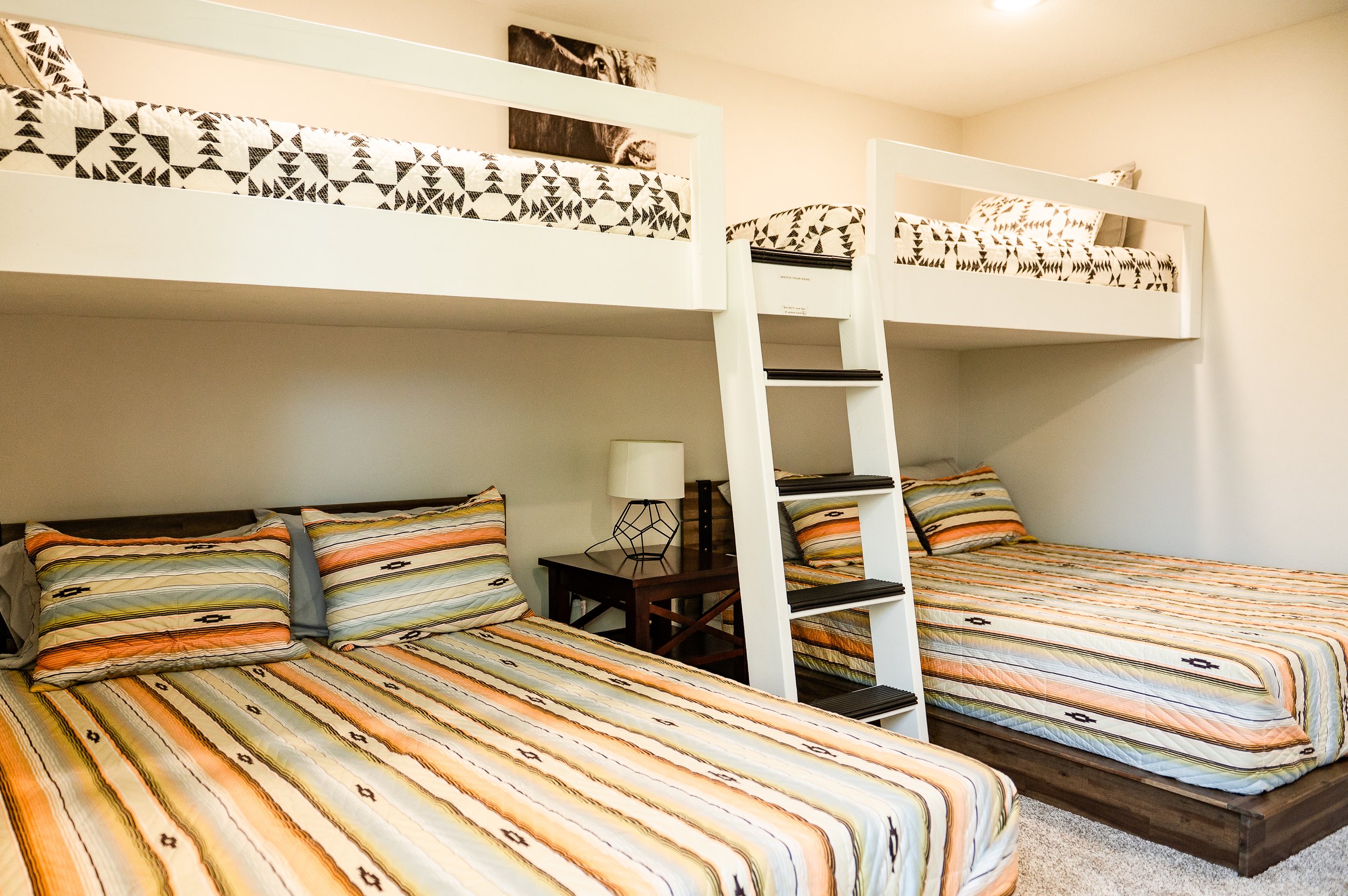 A bedroom with two full beds, each covered with striped comforters and matching pillows. There is a small nightstand with a modern lamp between the beds, and a bunk bed with a white frame and ladder. A black and white lion portrait hangs above the bu