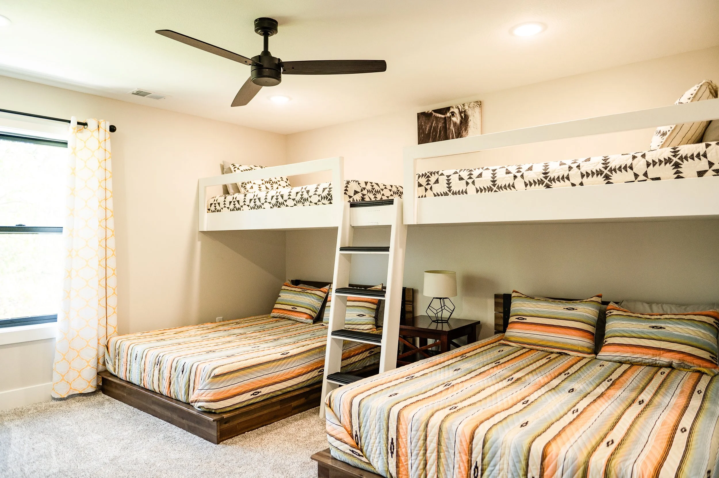 A bedroom with two beds, each with colorful striped bedding and pillows, a nightstand with a geometric lamp, a ladder leading to a top bunk with black-and-white tribal patterned bedding, a window with yellow patterned curtains, a ceiling fan, and a f