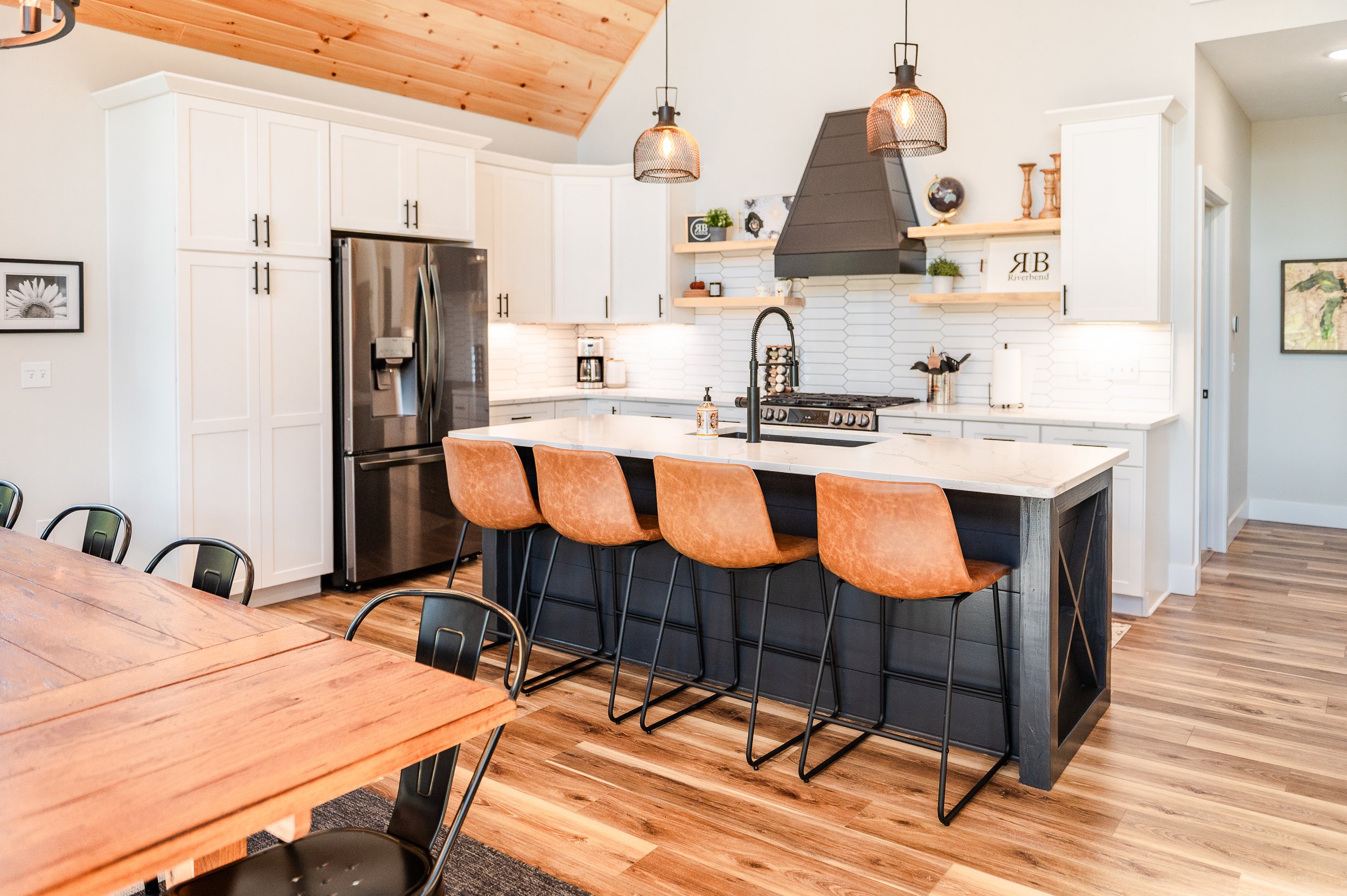 Modern kitchen with white cabinets, a black range hood, a marble island with orange chairs, and wood accents on ceiling and flooring.
