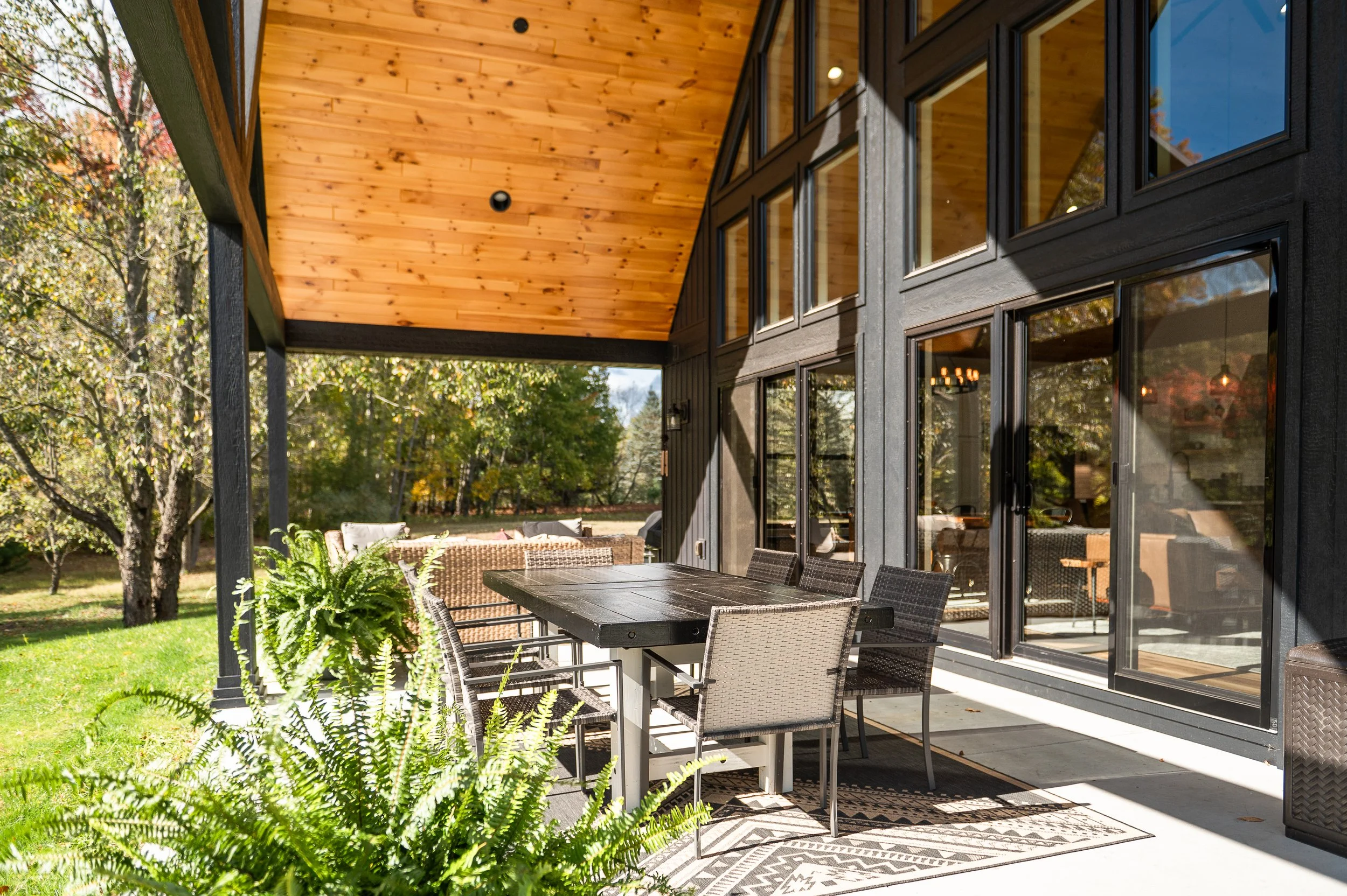 Outdoor patio with a black dining table and chairs, attached to a house with large glass windows, surrounded by trees with fall foliage.