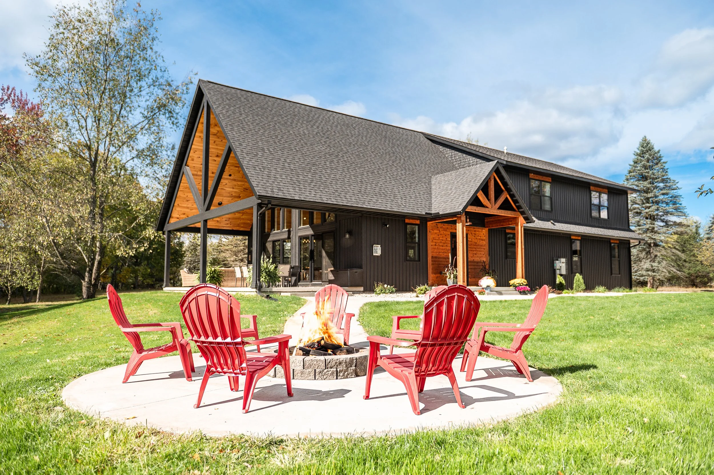 A backyard scene with a fire pit surrounded by red Adirondack chairs on a concrete patio. In the background, there is a modern black house with wood accents, large windows, and a covered porch, set against green trees and a partly cloudy sky.