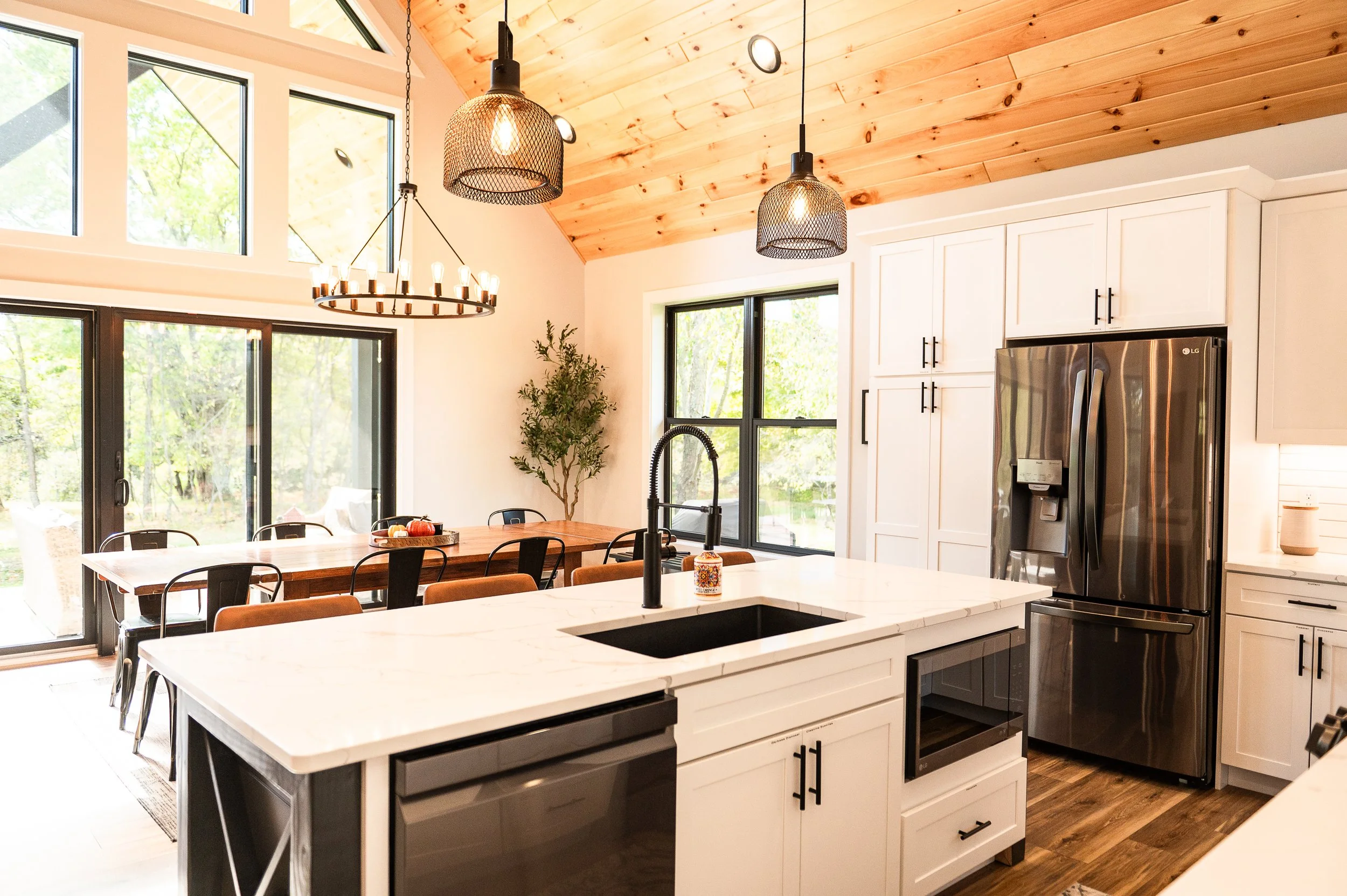Modern kitchen with white cabinetry, stainless steel refrigerator, black faucet, white marble countertops, large windows, wooden ceiling, pendant lights, and a dining area with a wooden table and black chairs.