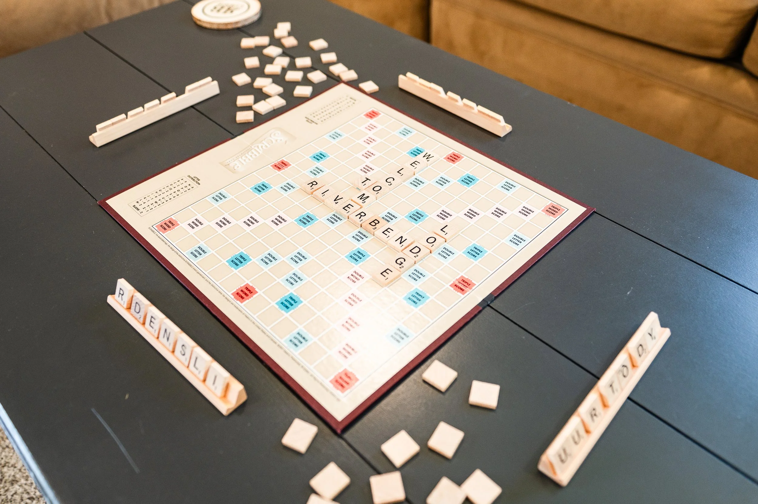 A Scrabble game on a black table showing the word 'RIVER' and 'BEND' in play, surrounded by Scrabble tiles and racks.
