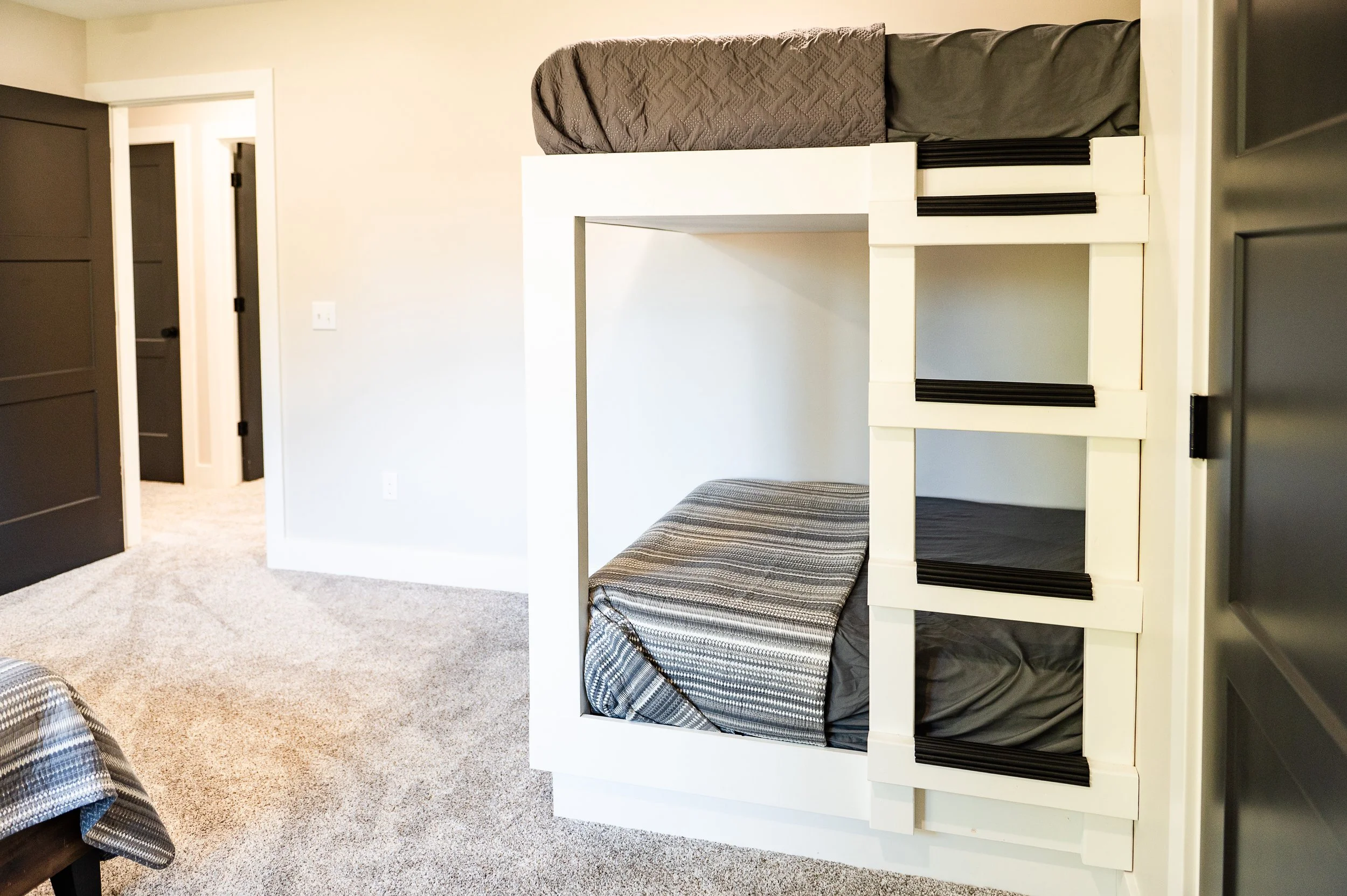 A bedroom with a bunk bed, with the lower bunk covered by a plaid blanket and the upper bunk with a gray blanket. A doorway leading to a hallway is visible in the background. The room has beige carpet and cream walls.