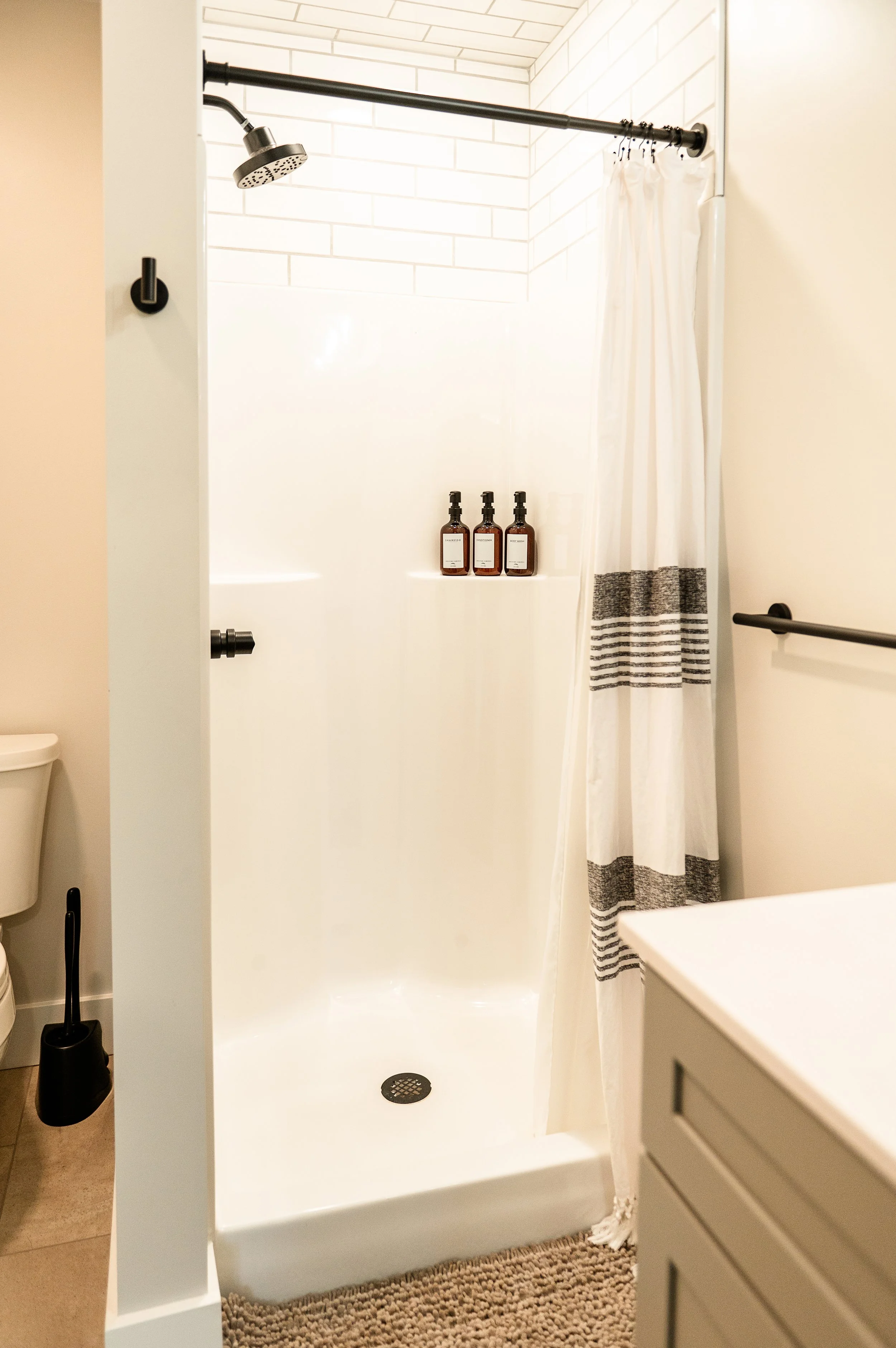 Bathroom shower area with a black shower head, a white shower curtain with black stripes, three brown bottles of soap or shampoo on a ledge, and a brown textured rug outside the shower.