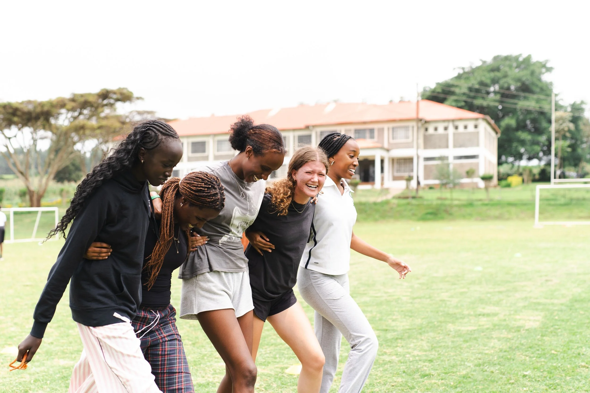Five women standing closely together outdoors on a grassy field, laughing and enjoying each other's company, with a large building and trees in the background.