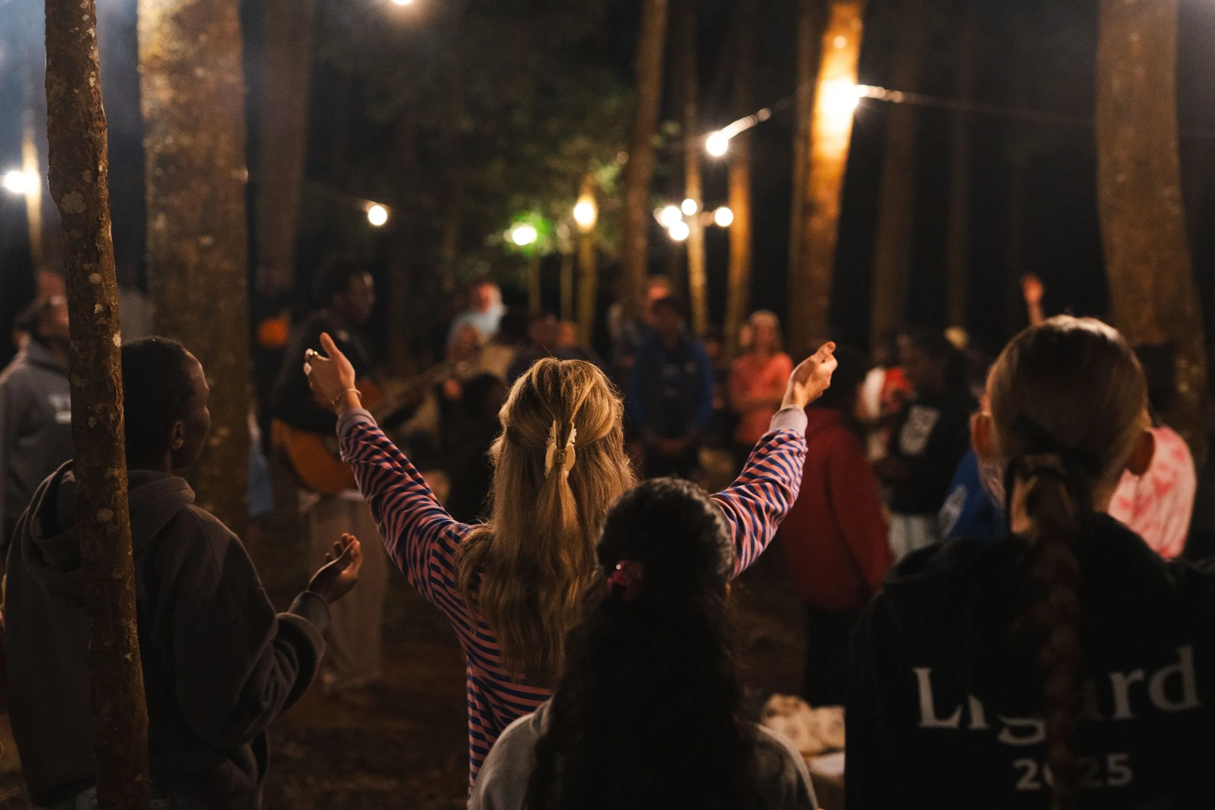 A group of people gathered outdoors at night, with string lights hanging among trees, as a woman in the center raises her arms in worship.