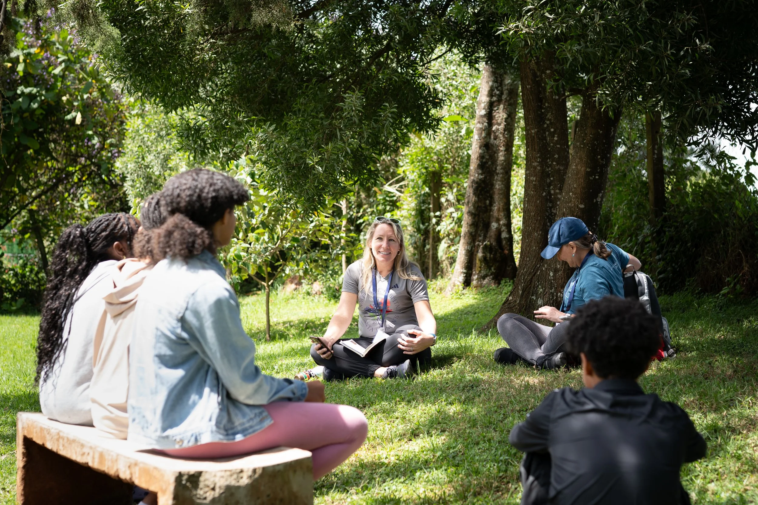 A diverse group of people sitting on the grass in a shaded park, listening to a woman speaking, who is holding a notebook, with trees in the background.