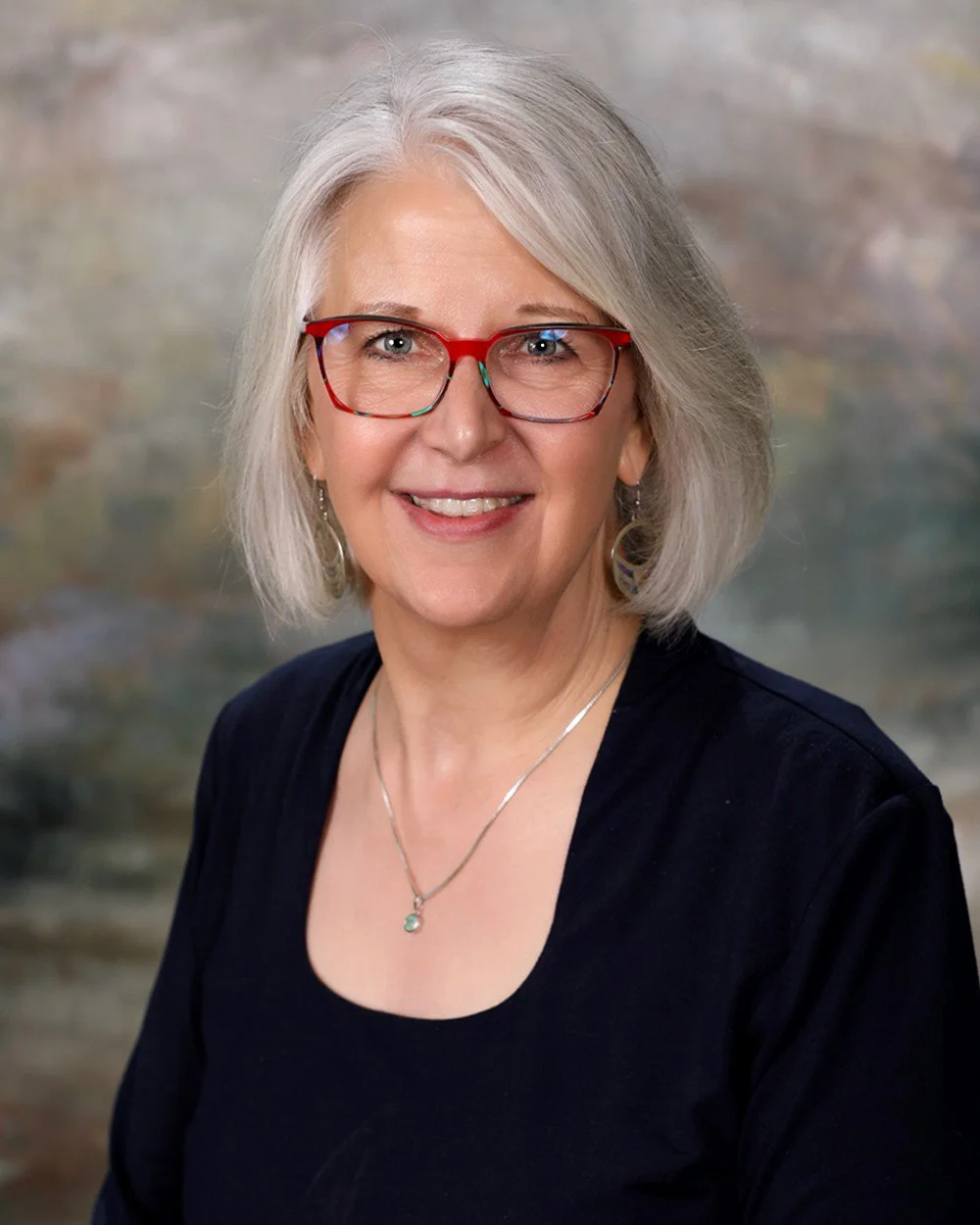 A smiling middle-aged woman with blonde hair and blue eyes, wearing a denim jacket and jewelry, standing in front of a rustic wooden background.