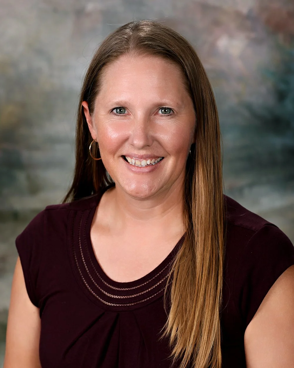 A woman with long brown hair, wearing a black top, smiling, with earrings and a necklace, standing in front of a white wooden background.