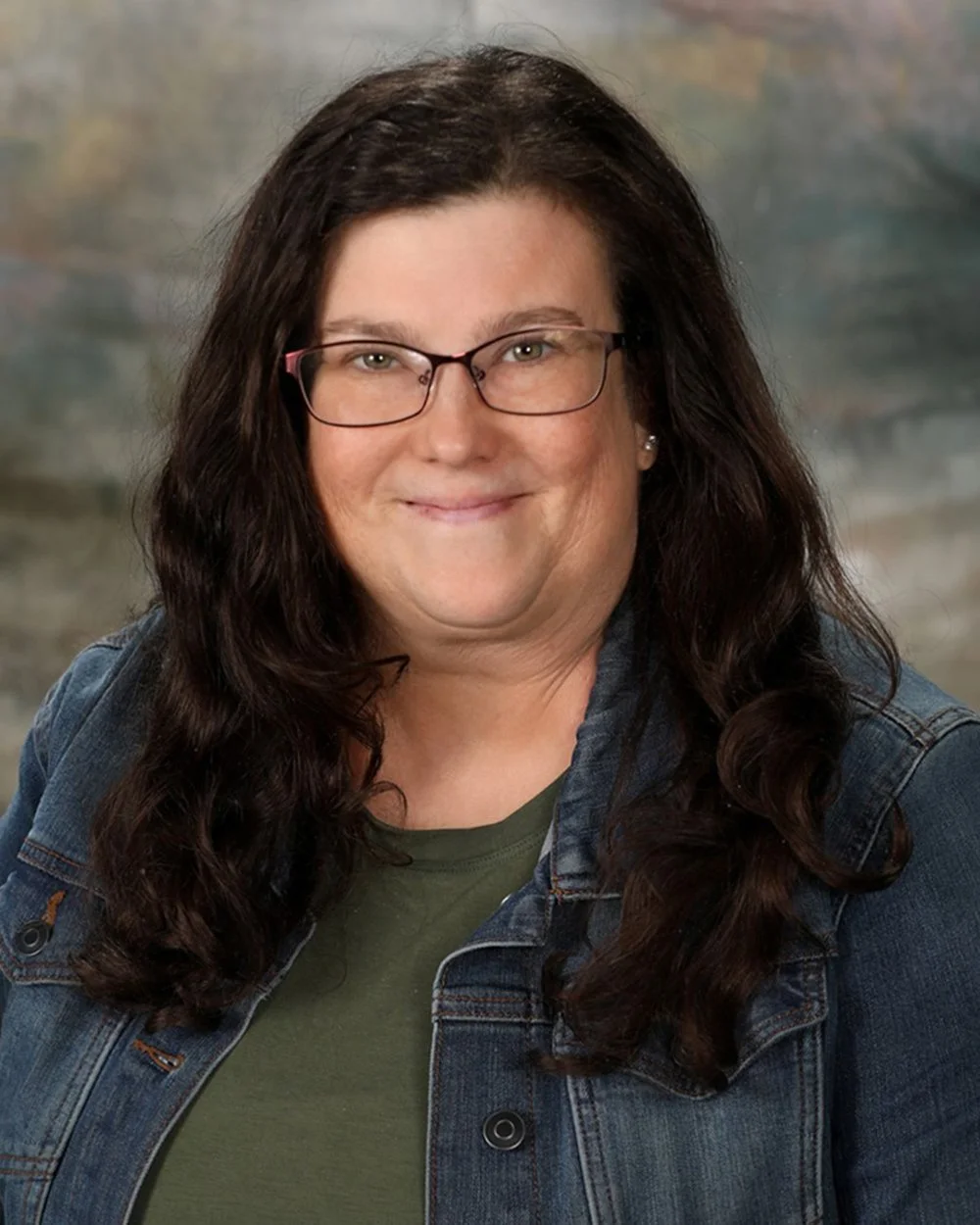 A woman with curly dark hair and glasses, wearing a gray patterned shirt and denim jacket, smiling at the camera in front of a light-colored background.