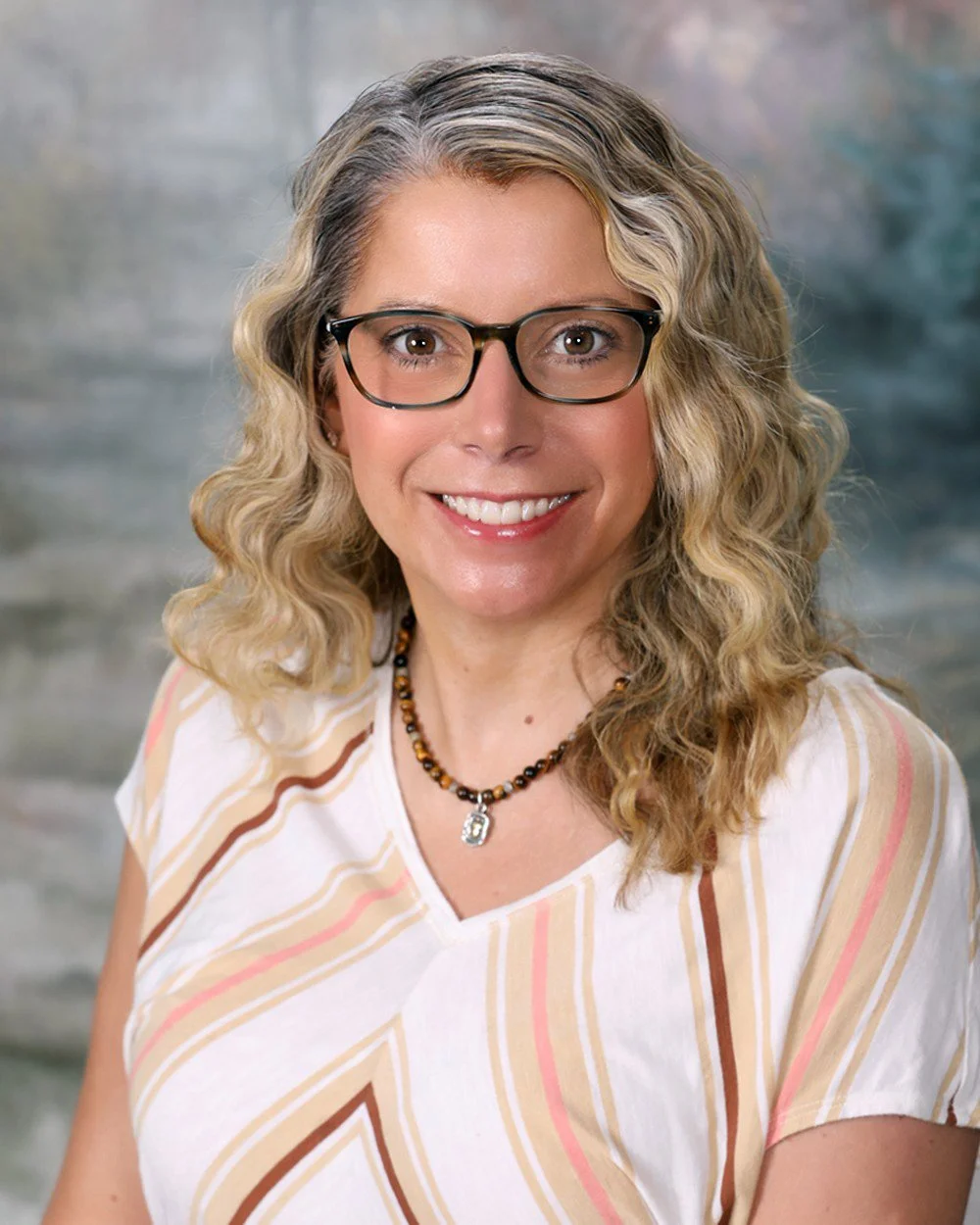 A woman with long wavy blonde hair, wearing glasses with a brown frame, a patterned blouse with pink and brown colors, a brown cardigan, and a necklace, smiling at the camera with a light-colored windowed wall in the background.