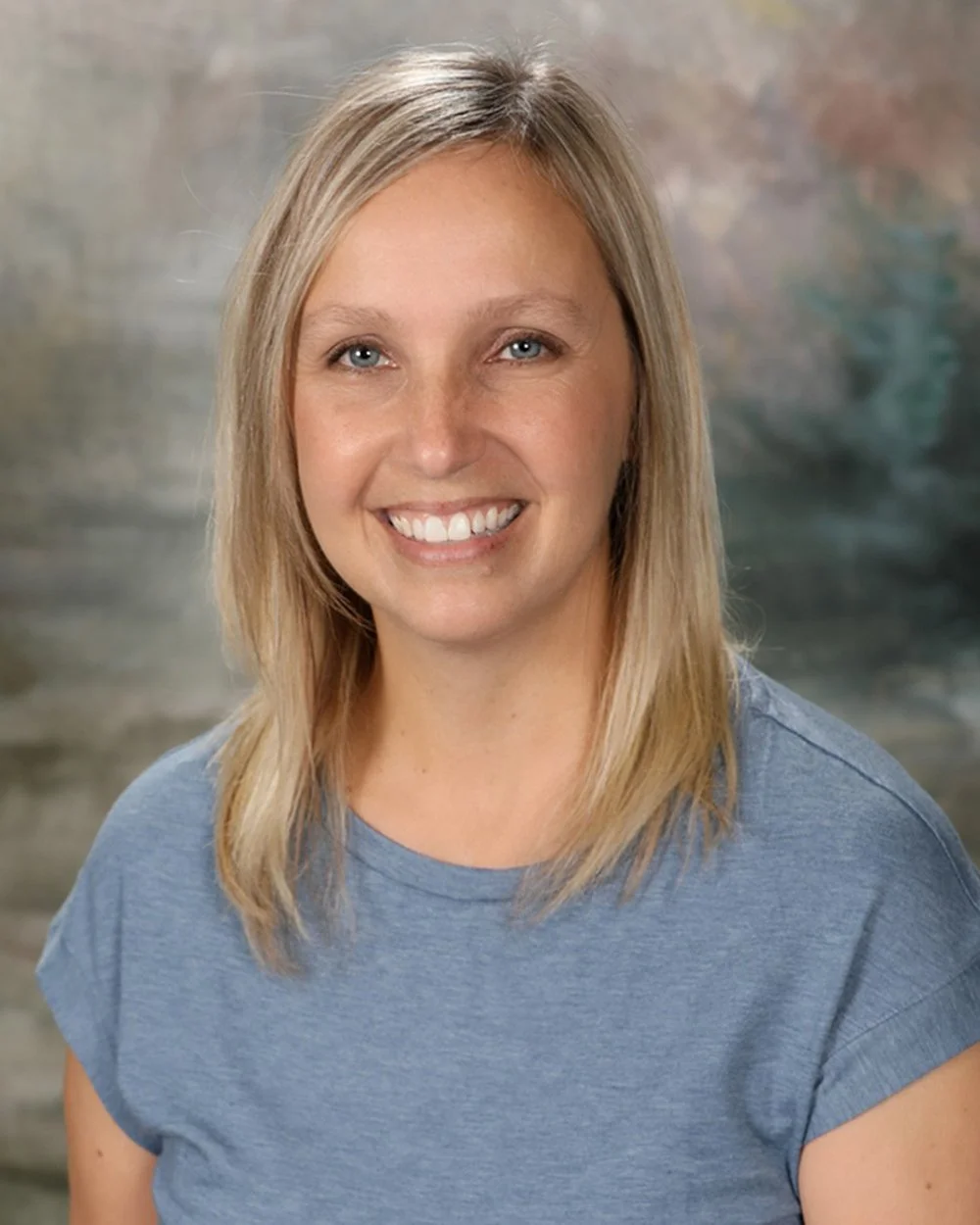 A woman with blonde hair and blue eyes smiling, wearing a dark green top, in front of a white panel background.