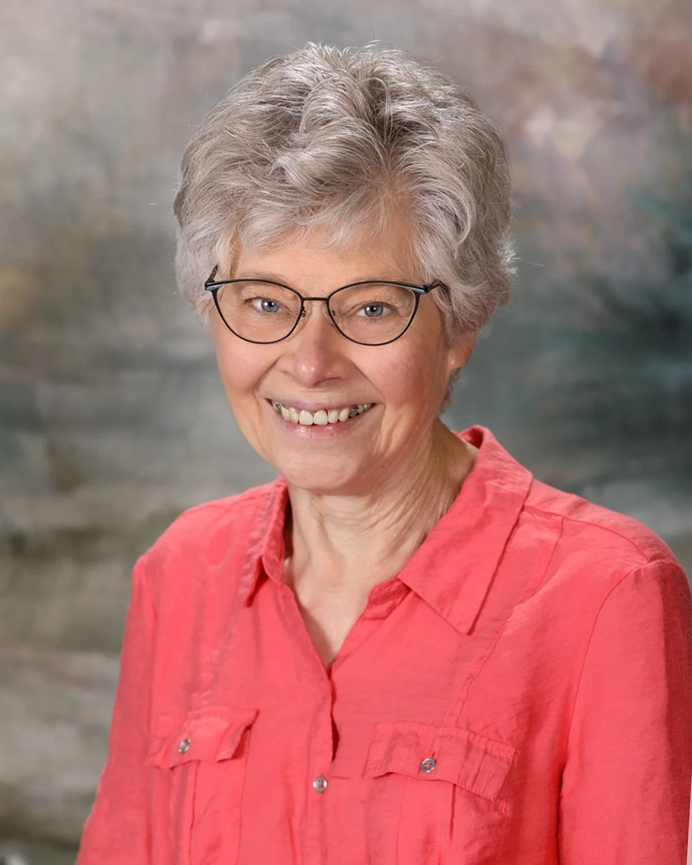 A woman with short gray hair, glasses, and a bright pink sweater smiling at the camera, standing in front of a wooden grid background.