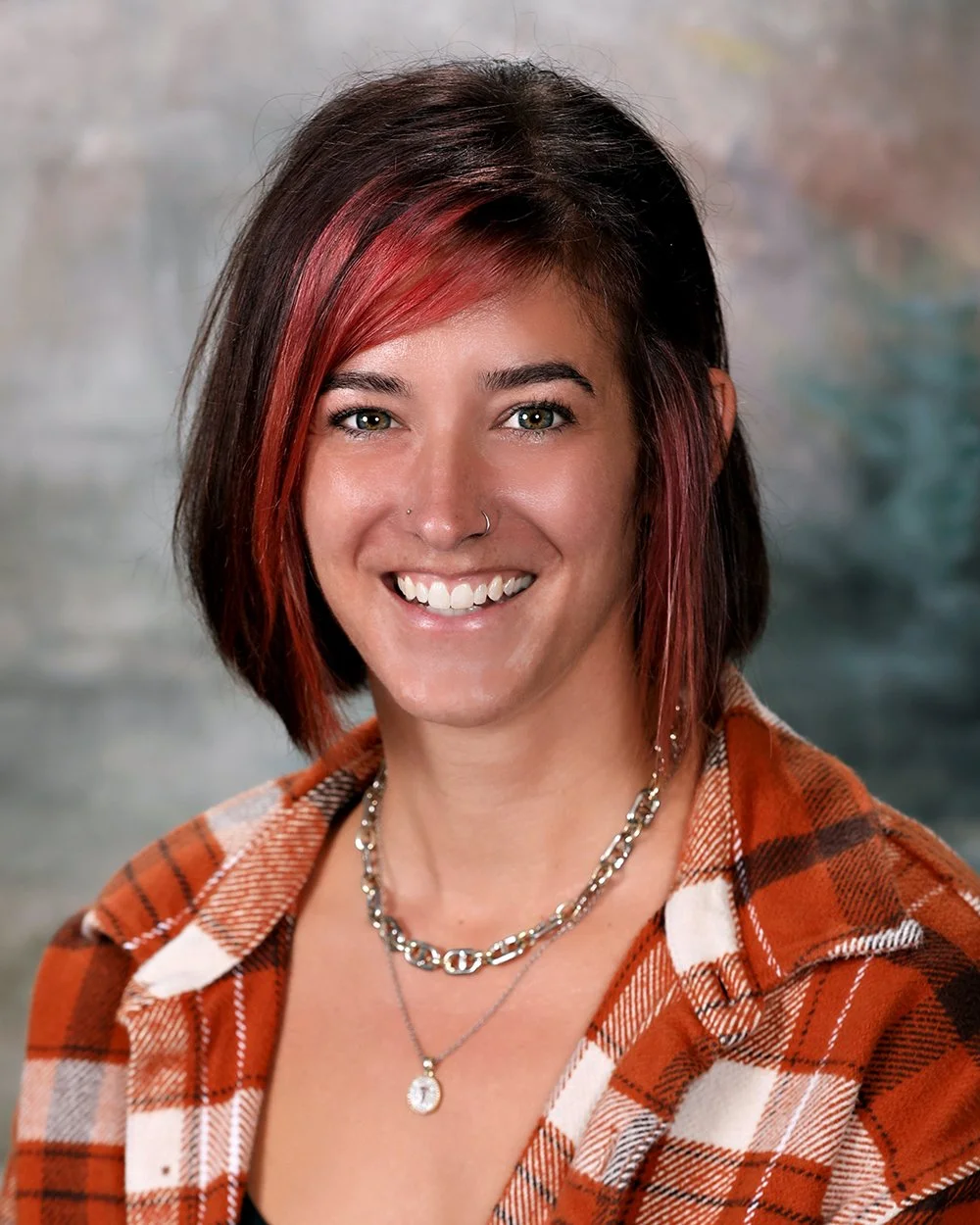 A woman with shoulder-length dark brown hair, wearing a black top and a green jacket with black leather sleeves, smiling at the camera.
